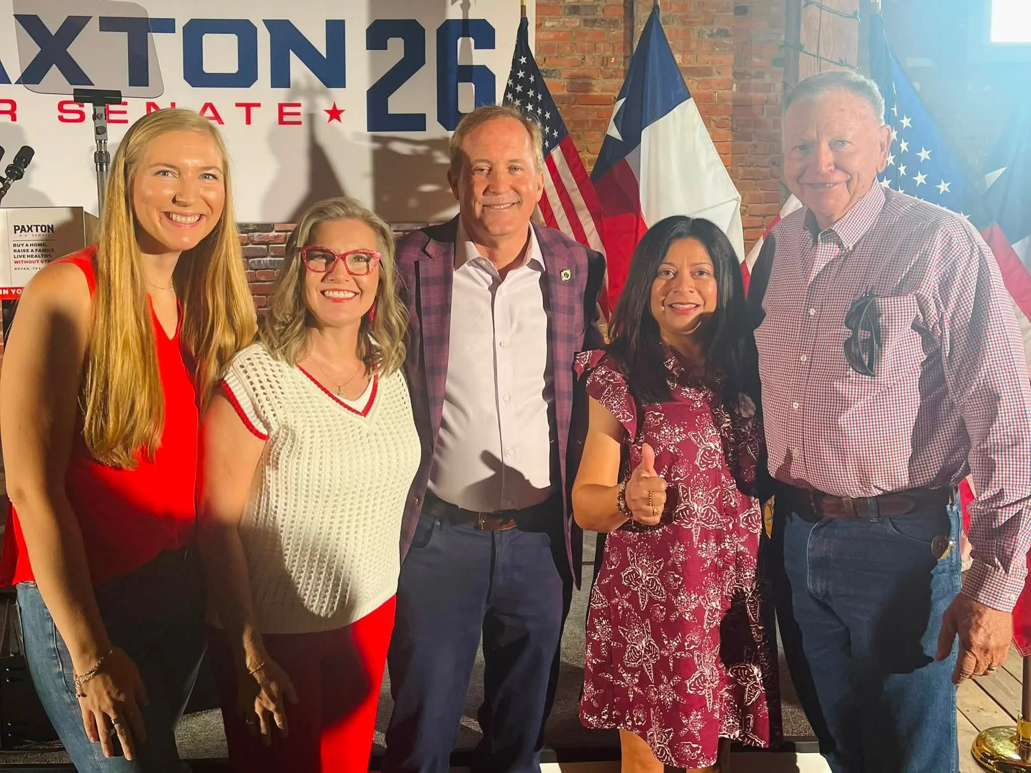 Group of people standing together at a political event, with American and Texas flags behind them, and a 'Paxton' campaign sign in the background.