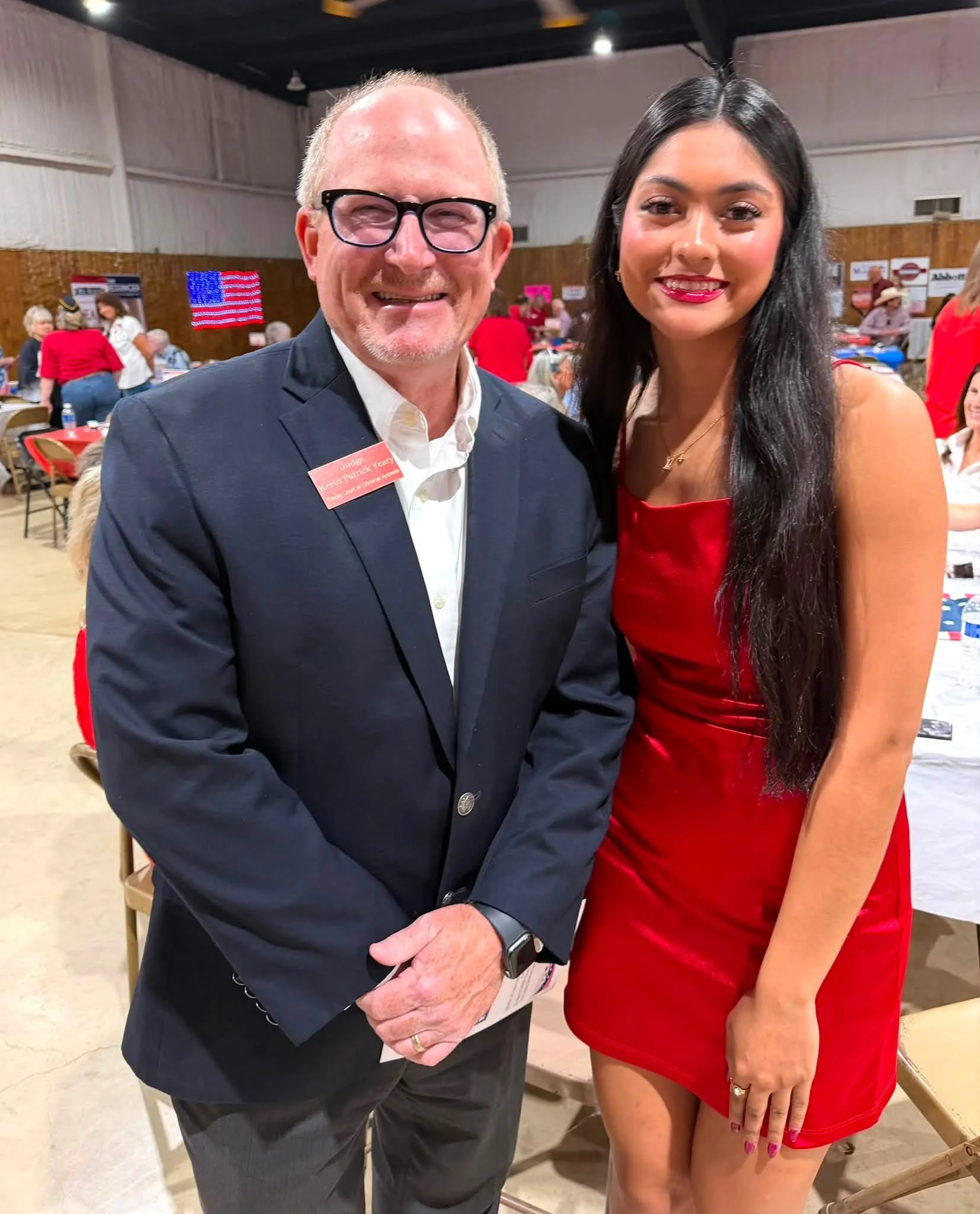 A man and a woman stand close together at an indoor event, smiling at the camera. The man is wearing a navy blazer, white shirt, glasses, and has a red name tag. The woman is dressed in a red dress and has long dark hair. There are tables and people 