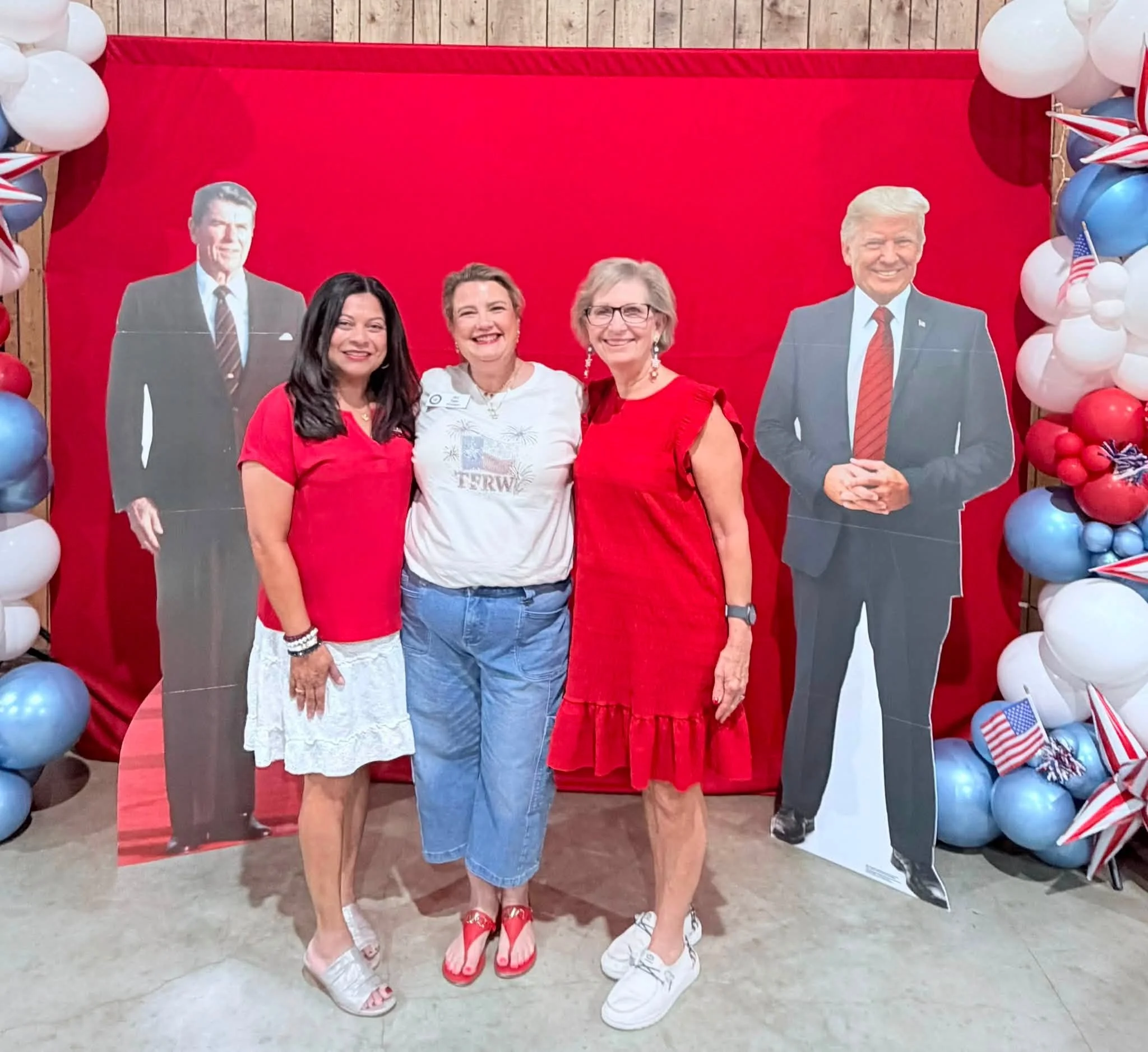 Four women standing together in front of a red backdrop decorated with patriotic balloons and stars, celebrating a patriotic event or holiday, smiling and dressed in red, white, and blue.