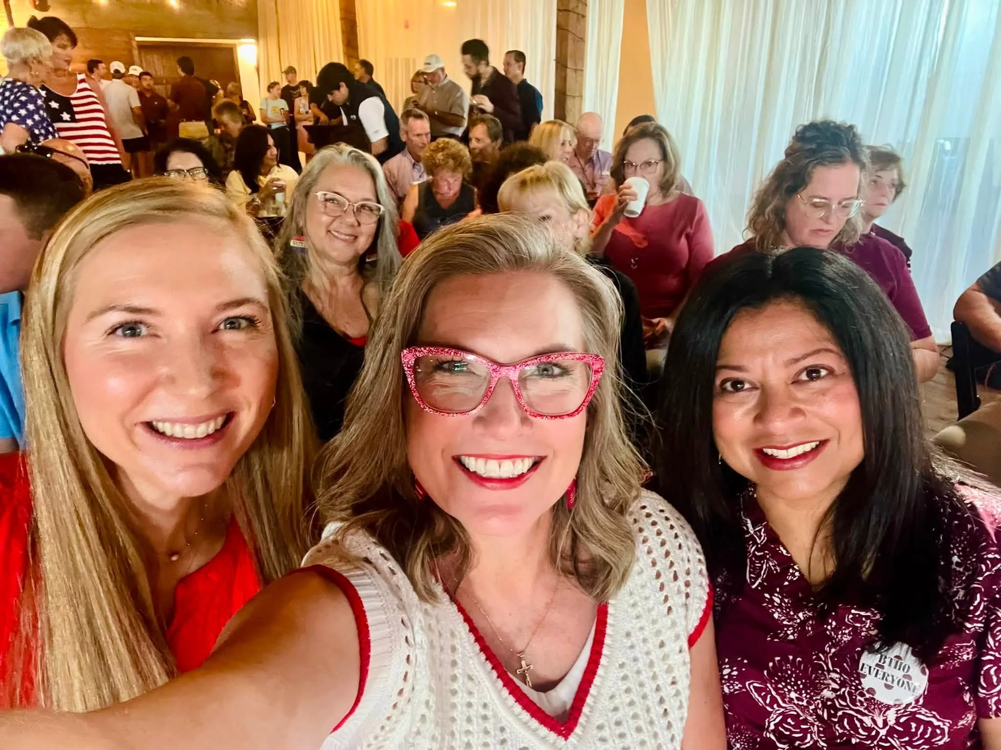 Group of people taking a selfie at an indoor event with wood-paneled walls and white curtains.