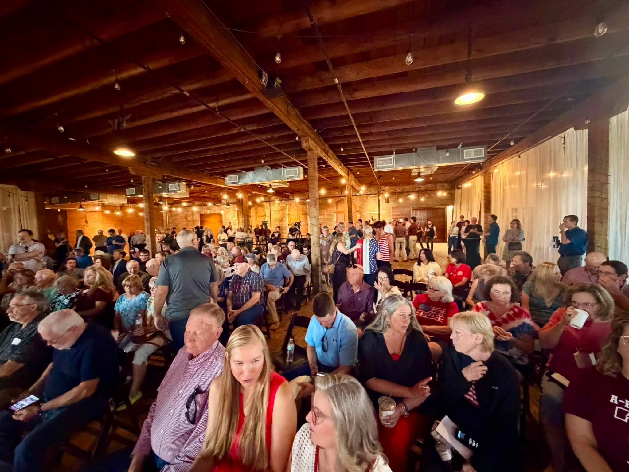 Crowd of people gathered in a large, rustic indoor space with a wooden ceiling and warm lighting, socializing and some using their phones.