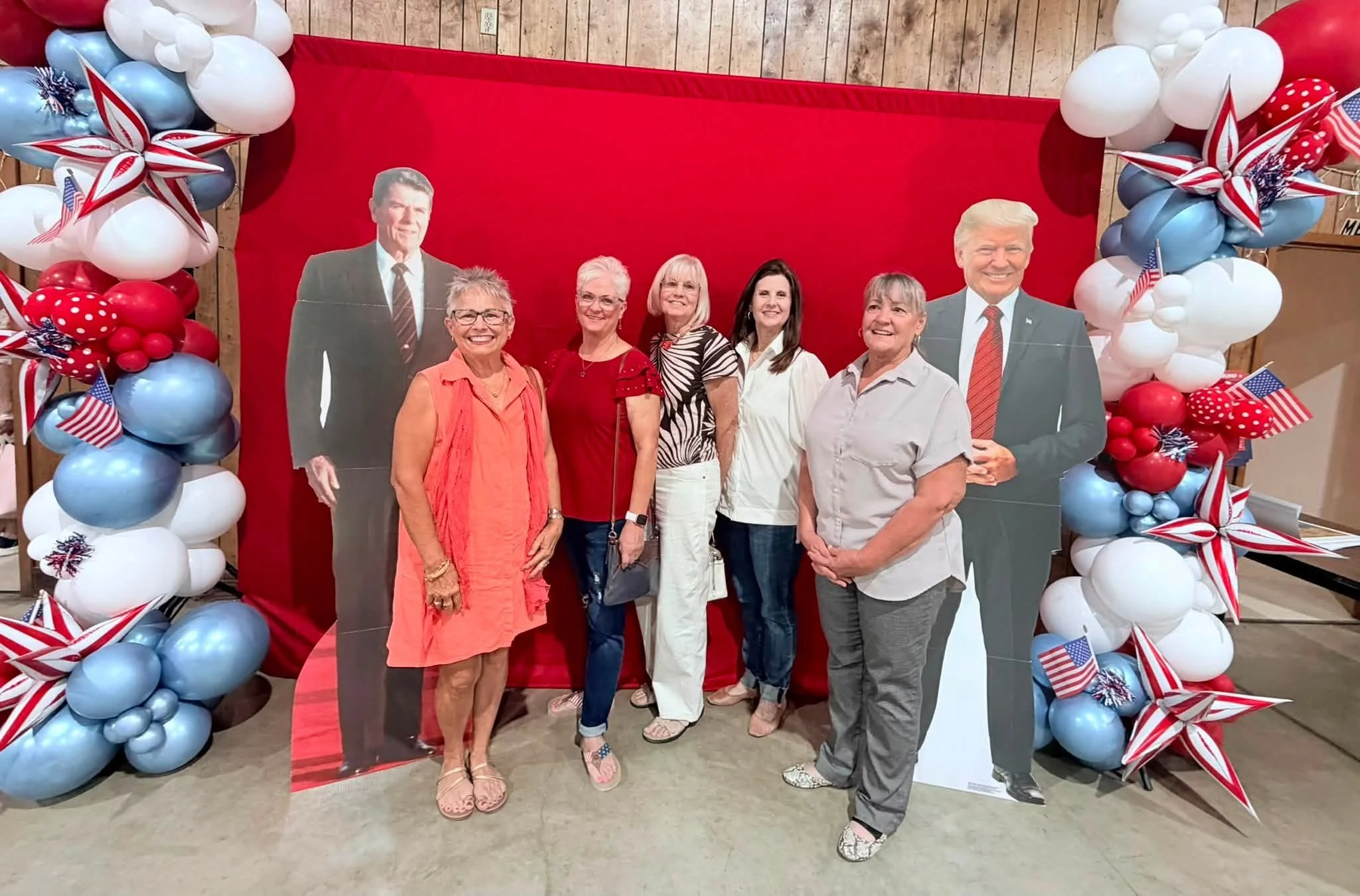 A group of six women standing in front of patriotic red, white, and blue balloon decorations, with cardboard cutouts of Donald Trump and another man, at a celebration or event.