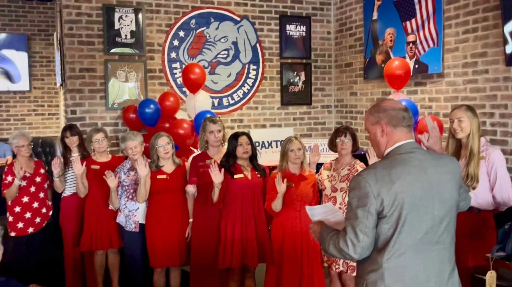 A group of women dressed in red, many with patriotic patterns or symbols, are participating in a ceremony in a room decorated with political posters, balloons, and banners. One man in a gray suit appears to be leading the event, while another woman i