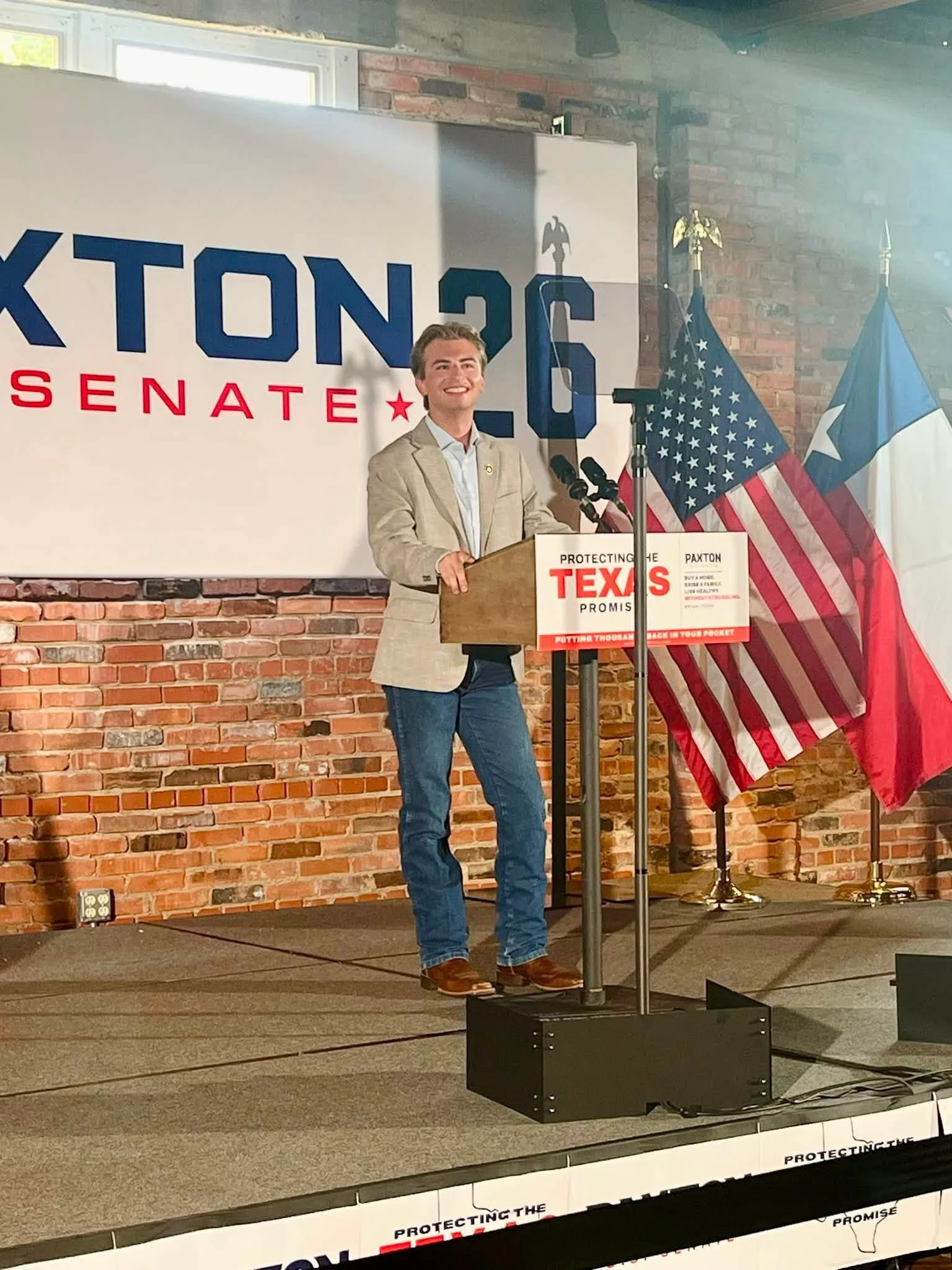 A man standing at a podium speaking at a campaign event with a large sign reading 'KTON 26 SENATE' in the background and several flags, including the American flag and Texas state flag, behind him.