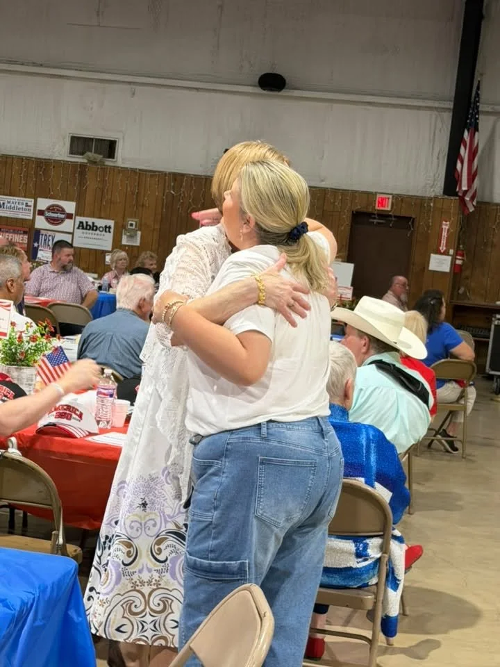 Two women hugging at a crowded indoor event with American flags and patriotic decorations.