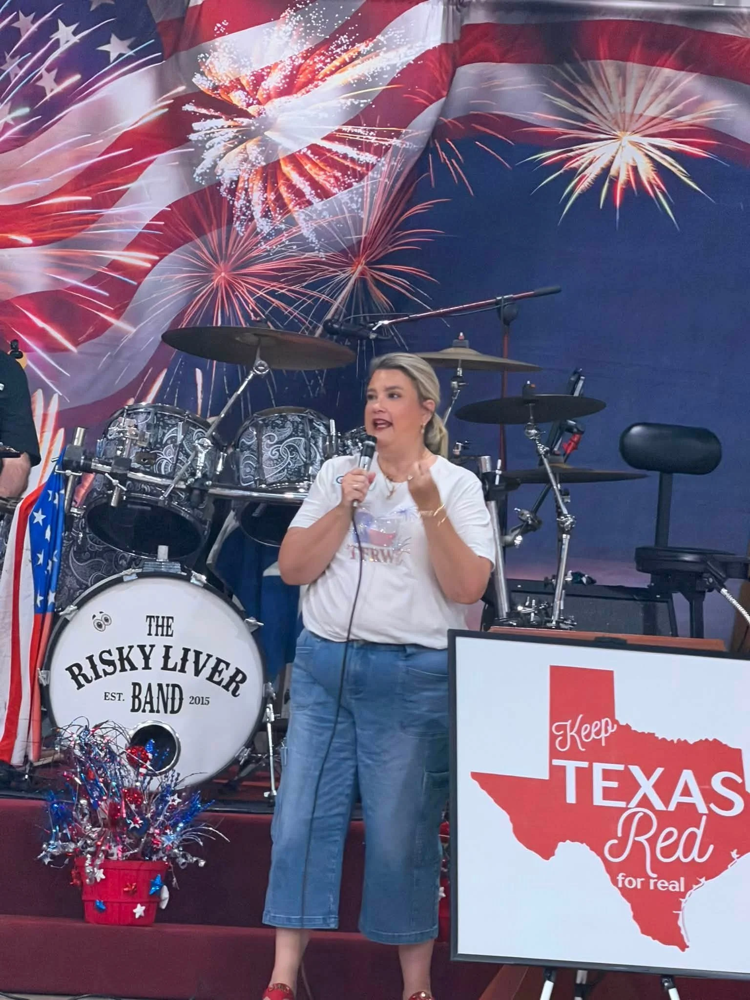 A woman holding a microphone speaks in front of a drum set with a white bass drum that reads 'The Risky Liver Band Est. 2015.' Behind her is a large American flag-themed backdrop with fireworks imagery. There is a red bucket filled with patriotic dec
