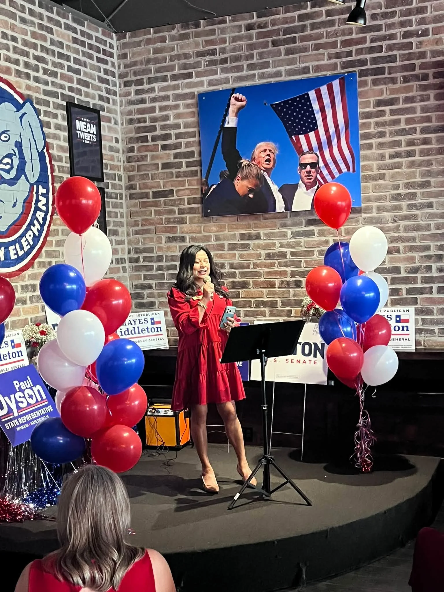 A woman in a red dress speaking into a microphone on a stage decorated with red, white, and blue balloons, with a brick wall background and political campaign signs. A large political poster behind her features a man and a woman with American flags.
