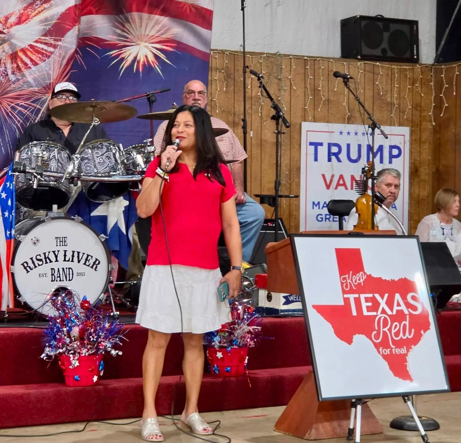 A woman in a red top and white skirt holding a microphone stands on stage, with a band behind her including a drummer and a man with a violin, as well as patriotic decorations and signs supporting Trump and Texas