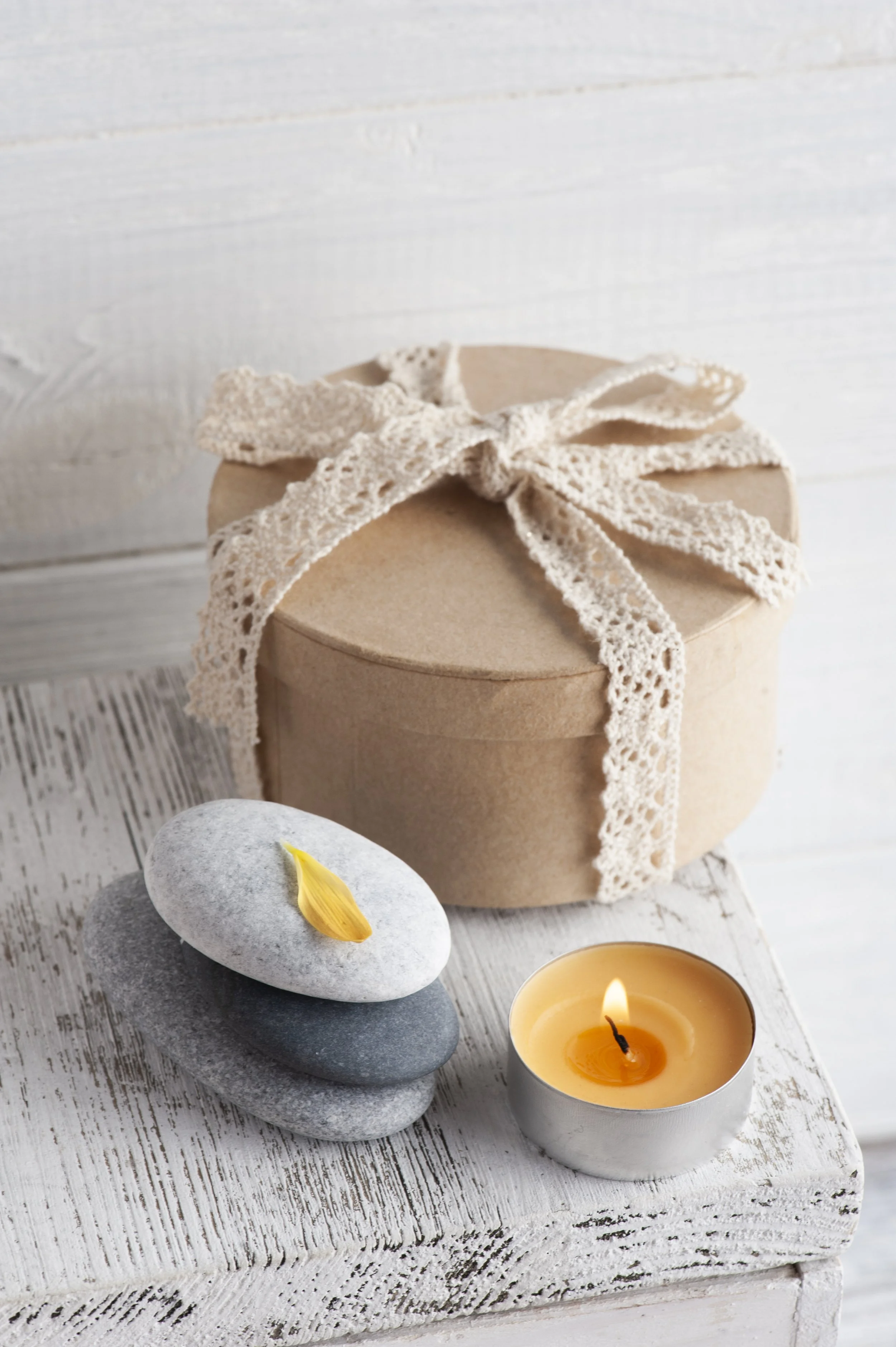 A round gift box with a lace ribbon, stacked stones with a flower petal, a lit candle, and a white wooden surface