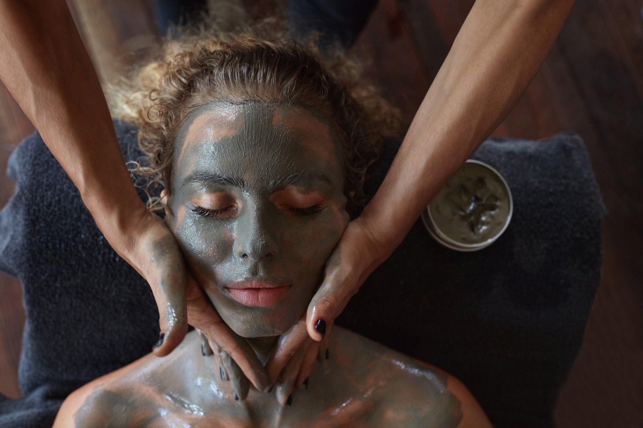 A person lying down with their eyes closed, receiving a facial treatment. A therapist applies a green clay face mask and the person appears relaxed. A bowl of the facial mask is visible nearby.