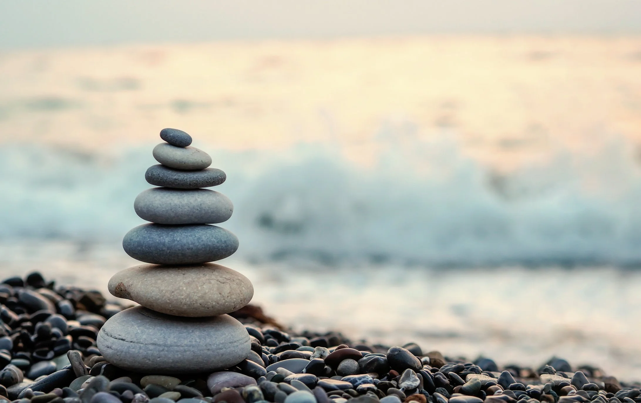 Stack of seven smooth, flat stones on a pebble-covered beach with the ocean and a cloudy sky in the background.