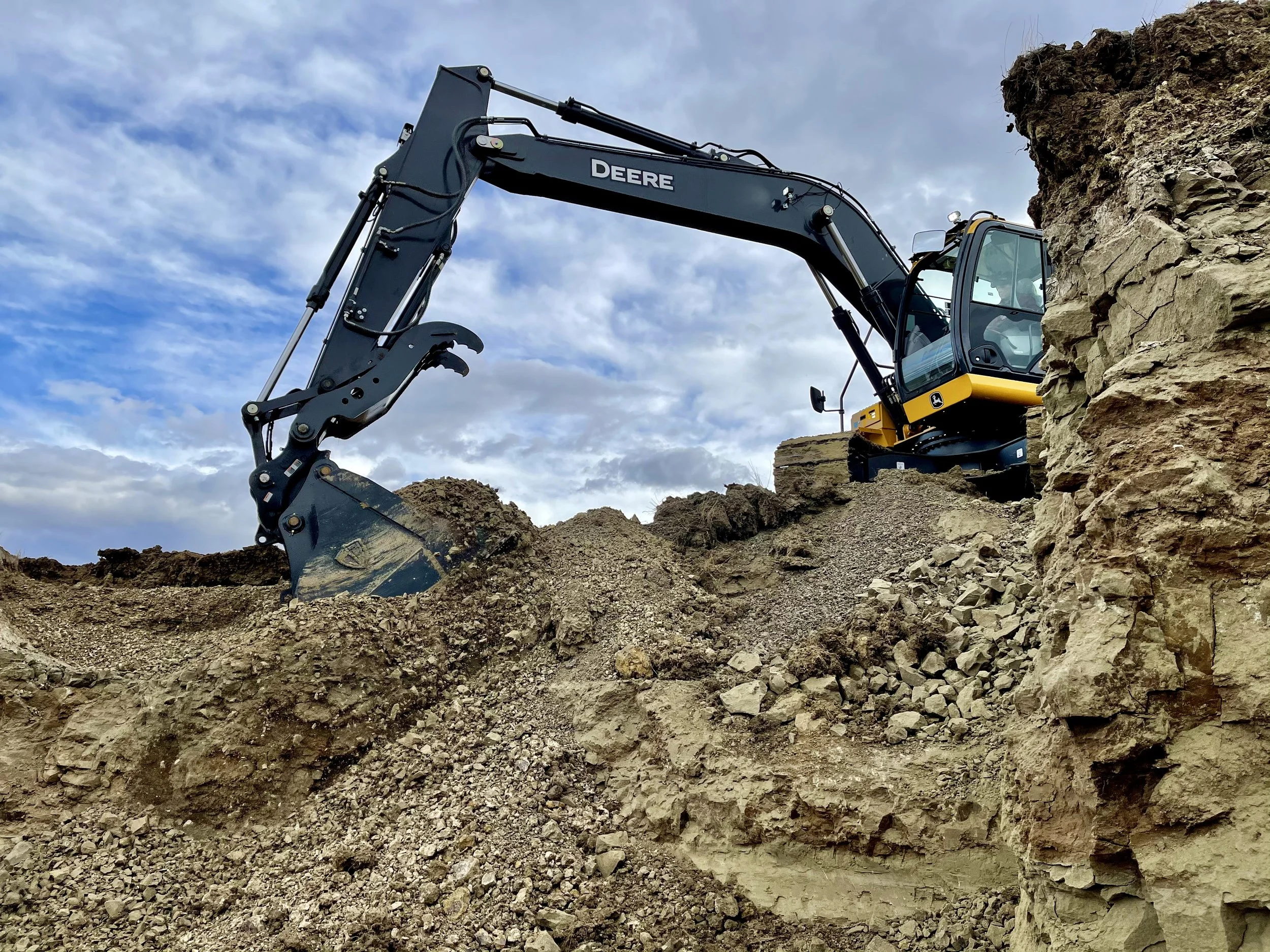 A John Deere excavator moving dirt and rocks in a construction or excavation site under a partly cloudy sky.