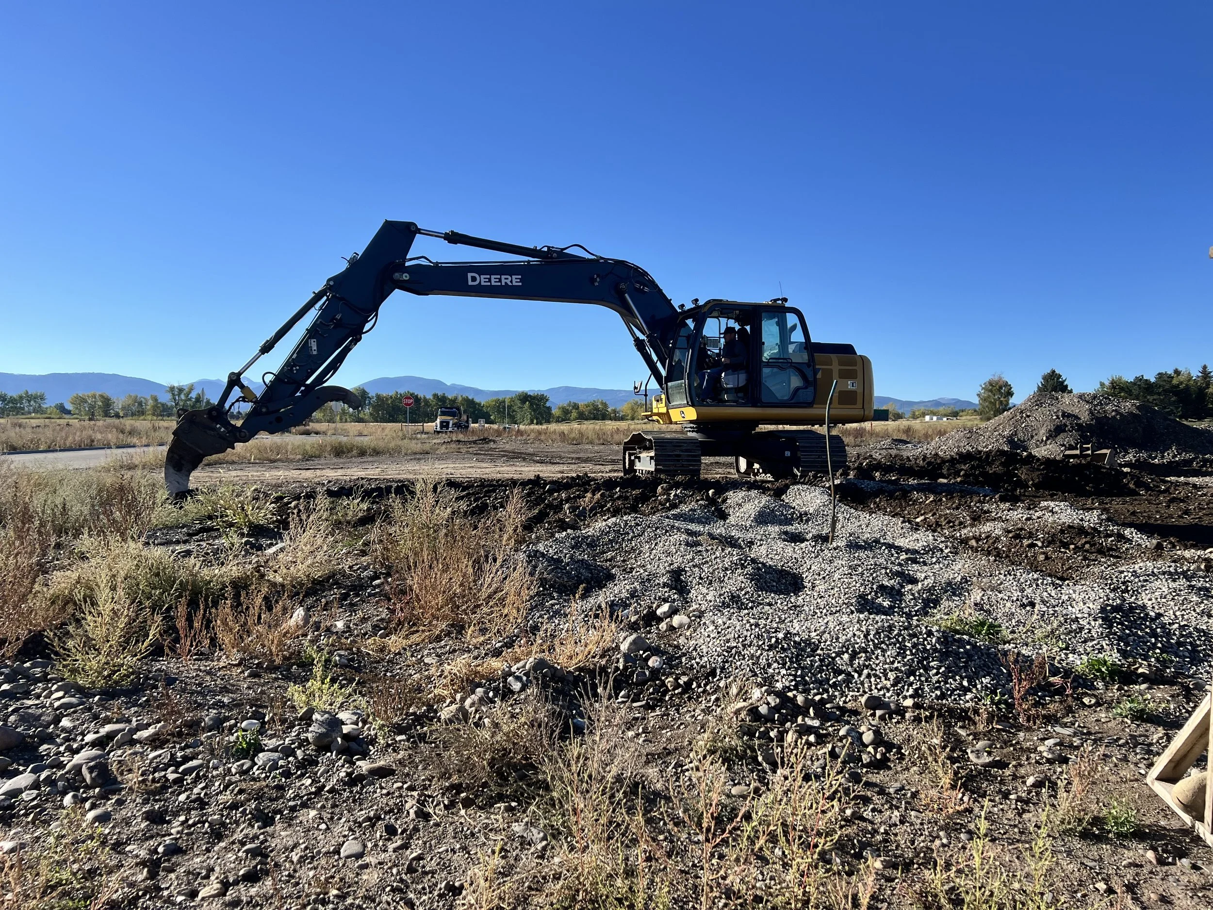 A John Deere excavator operating on a construction site with mountains in the background under a clear blue sky.
