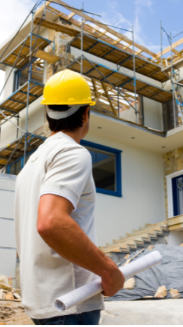 Construction worker holding blueprints at a building site with scaffolding and an unfinished house in the background.