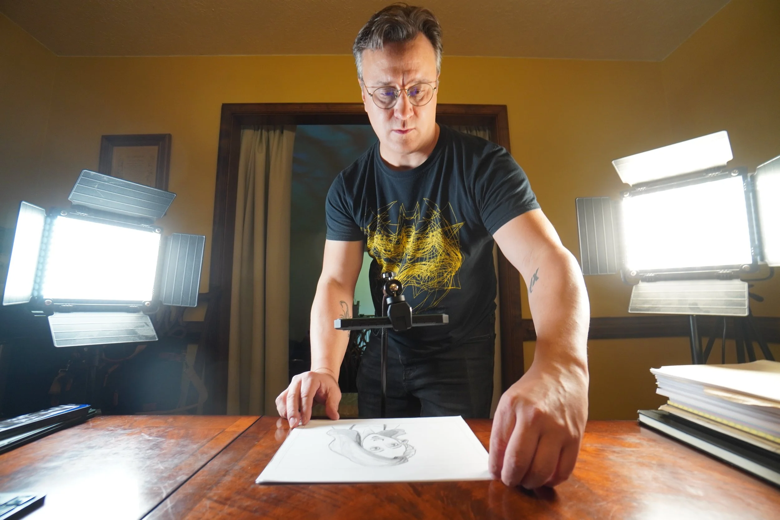 A man wearing glasses and a black T-shirt leans over a drawing of a woman's face on a white sheet of paper on a wooden table, with studio lights illuminating the workspace.