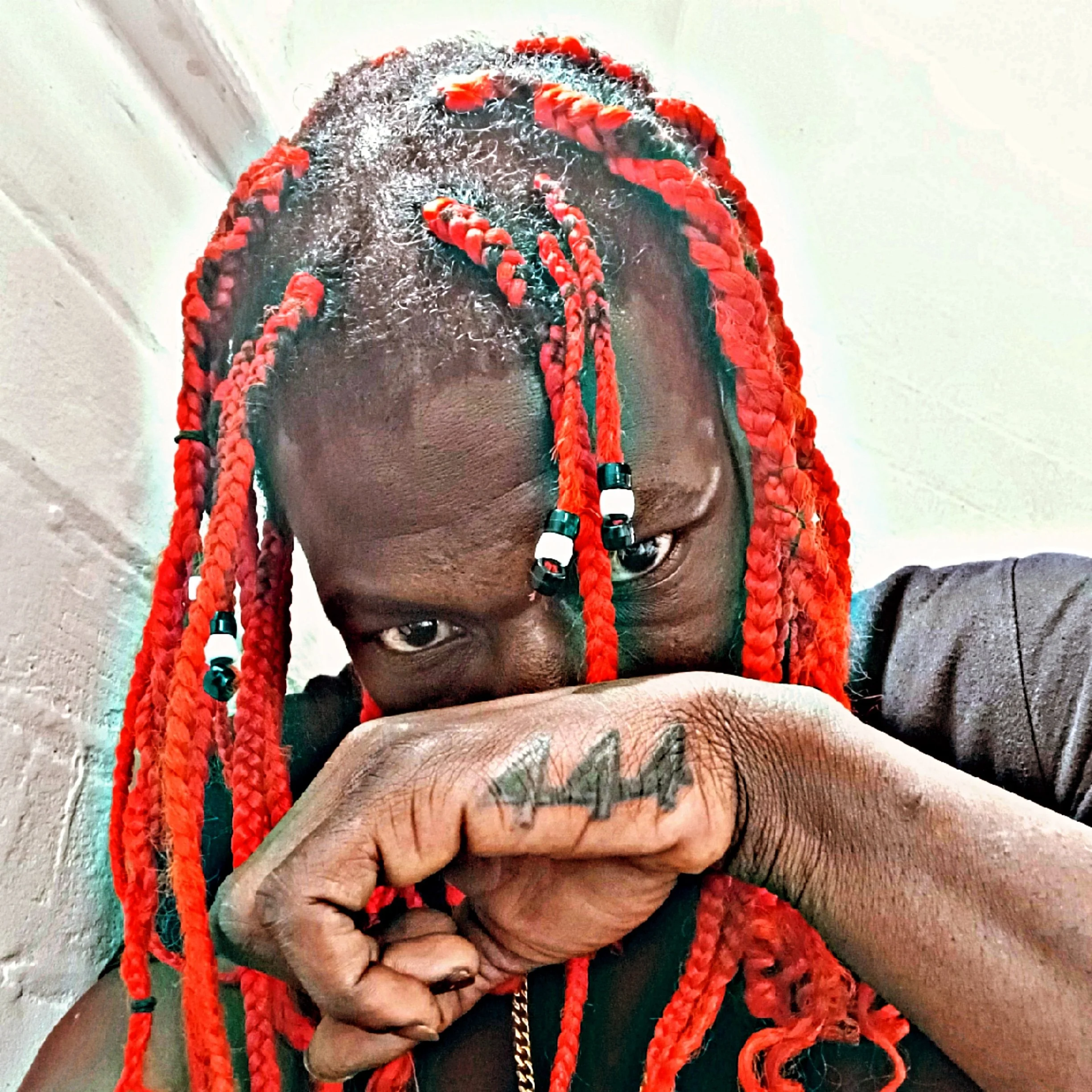 A person with braided red hair and dark skin, holding their fist up near their face. The person is wearing a dark grey shirt and has rings on their fingers, with a tattoo visible on their hand. The background appears to be a plain wall.