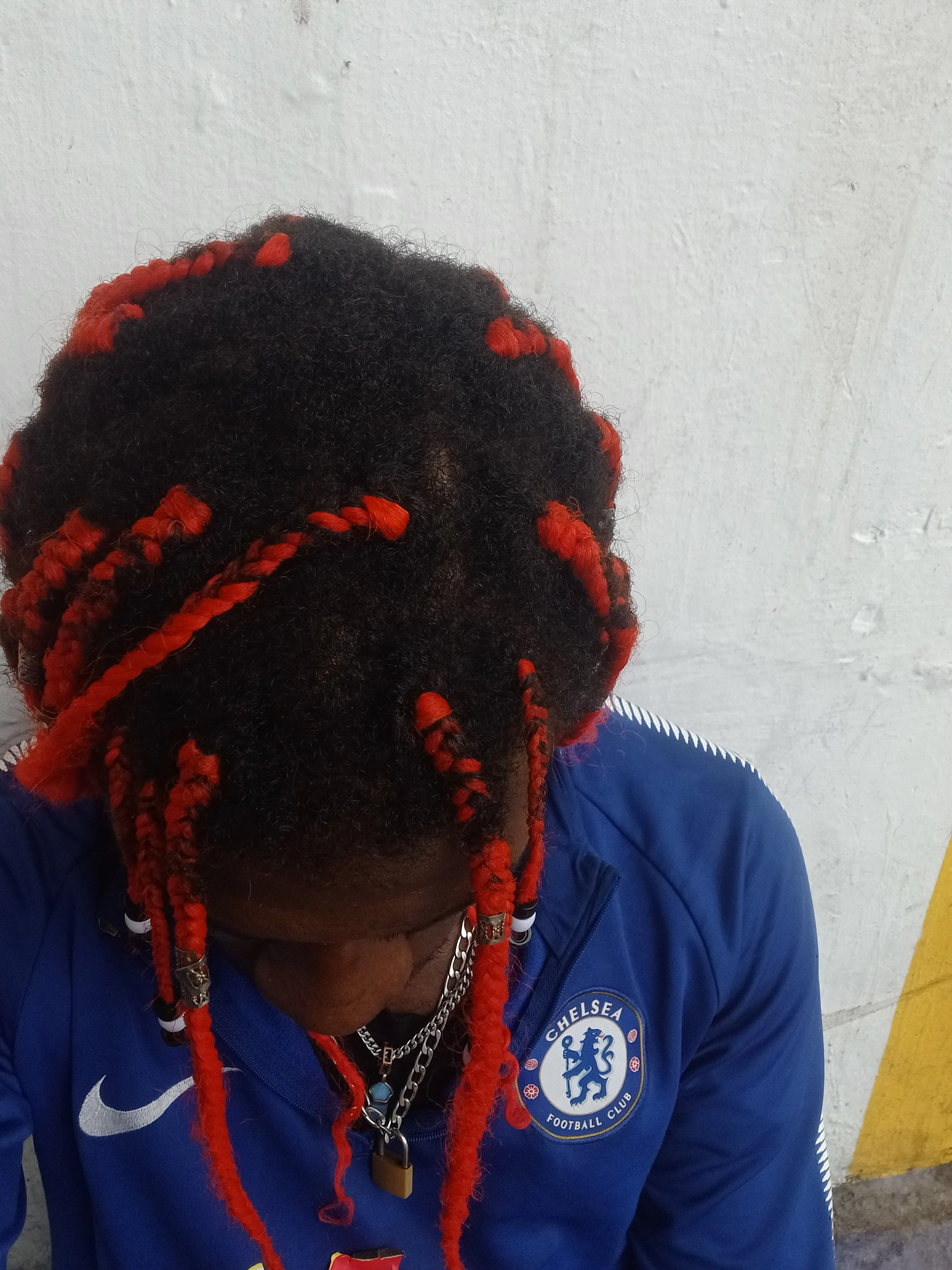 Close-up of a person with red and black hair styled in dreadlocks, wearing glasses, a blue Chelsea football club jacket, and multiple necklaces, against a plain white wall.