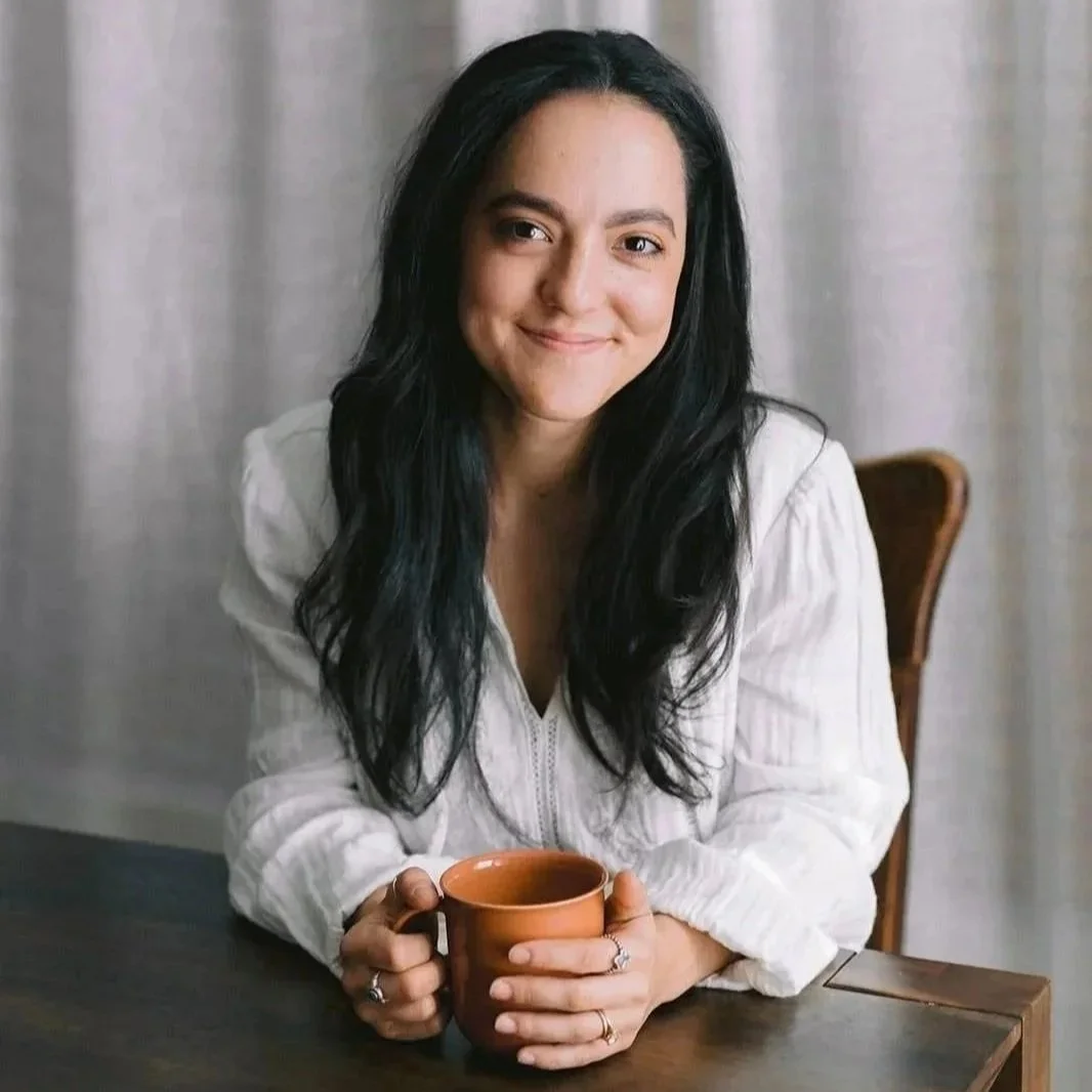 A woman with long black hair sitting at a table, holding a brown mug, smiling at the camera.
