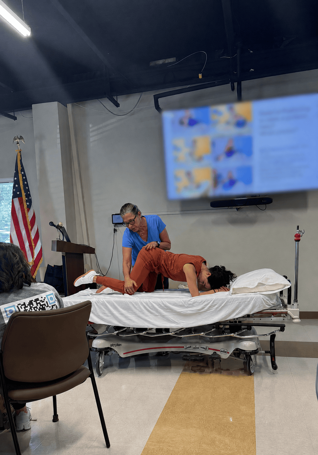 A woman performing a physical therapy move on a patient lying face down on a hospital bed in a clinical setting with an American flag in the background.
