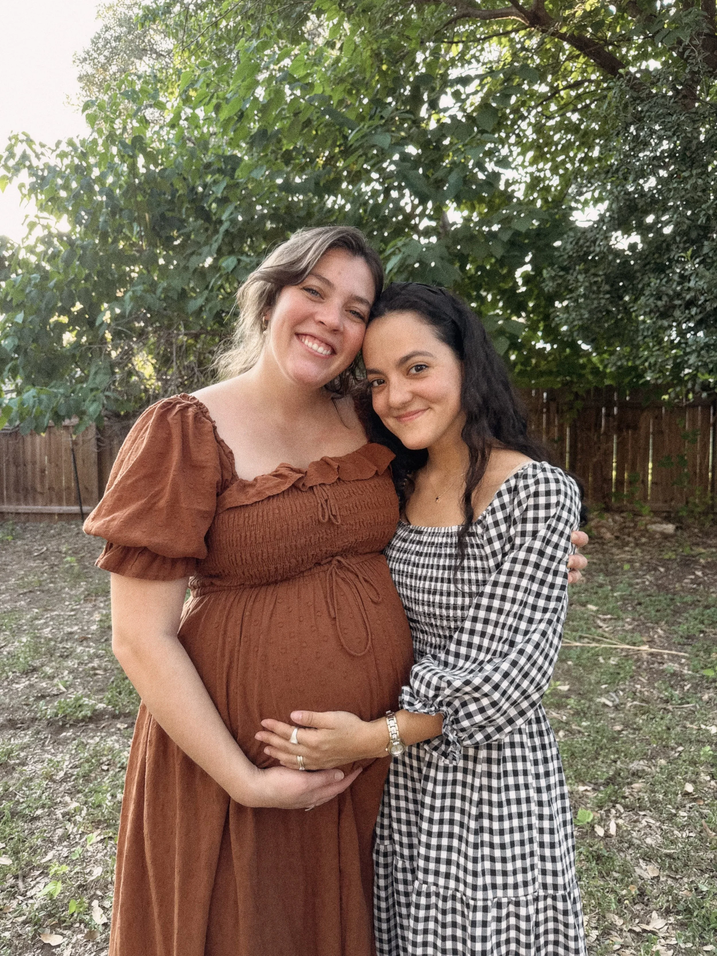Two women standing close together outdoors, smiling, with a wooden fence and green trees in the background. One woman is pregnant and the other is touching her belly.
