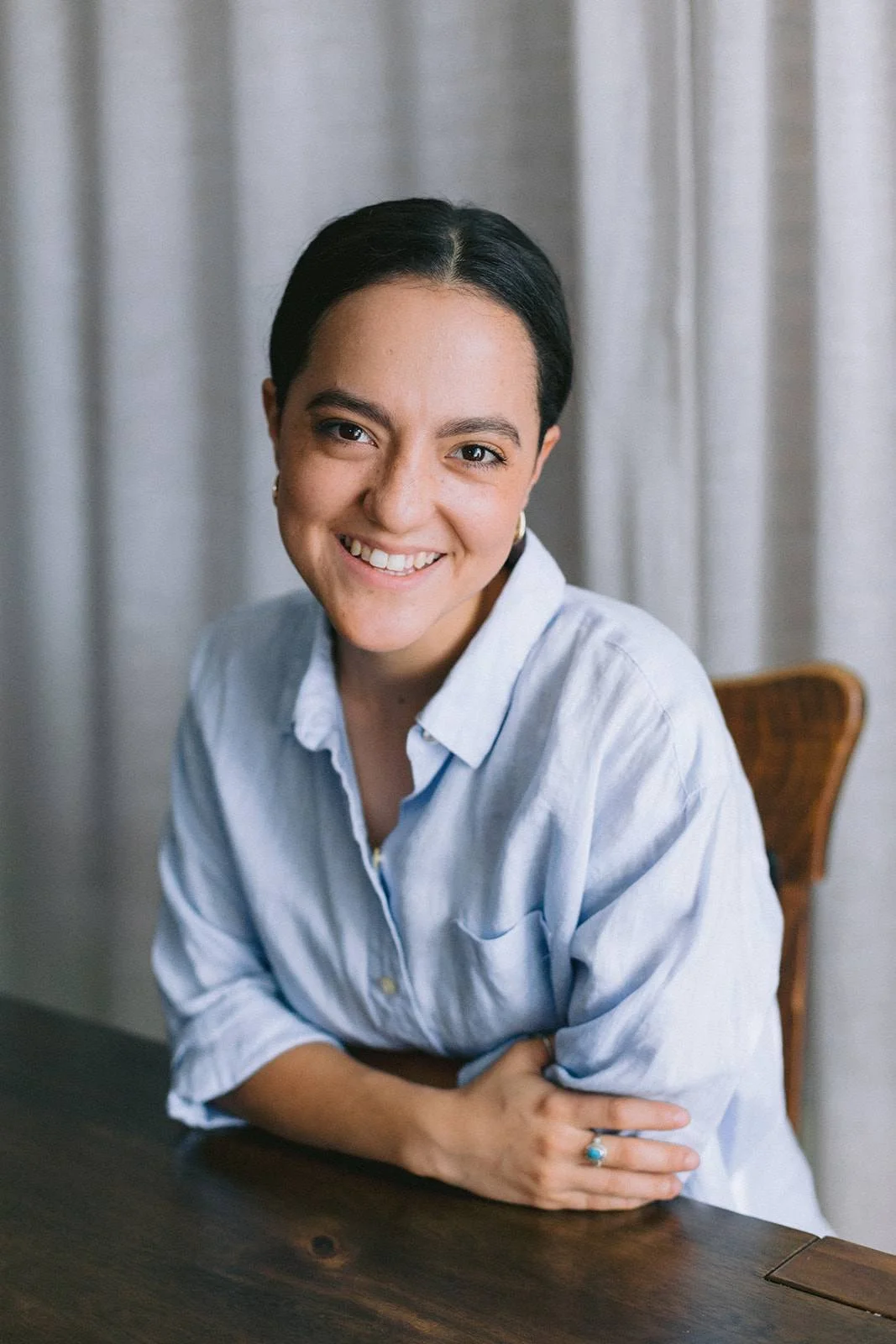 A woman with short dark hair and light skin, smiling, wearing a light blue button-up shirt, sitting at a wooden table with a beige curtain background.