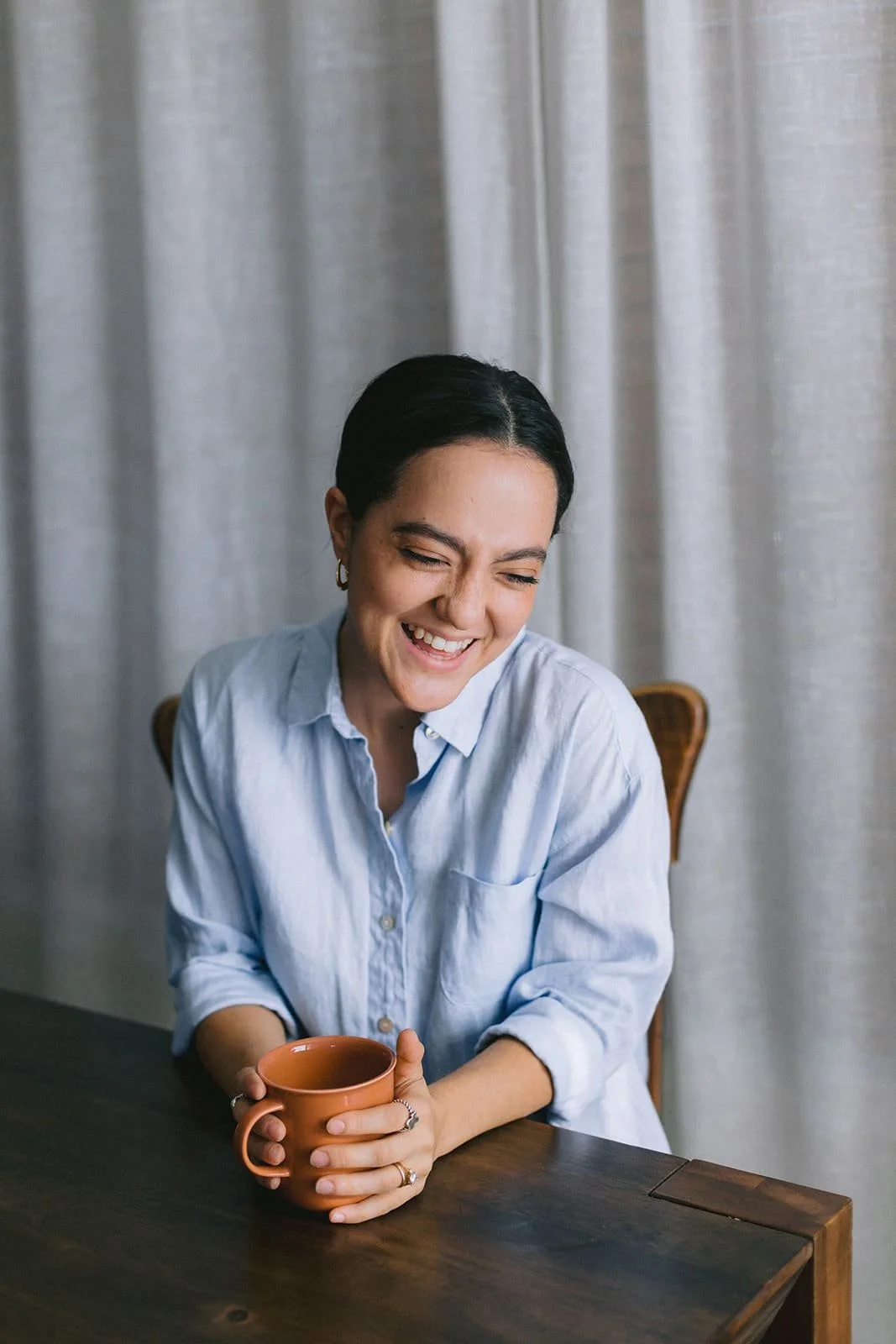 A woman with short dark hair, wearing a light blue button-up shirt, is sitting at a wooden table holding an orange mug, smiling and laughing with her eyes closed, in front of beige curtains.
