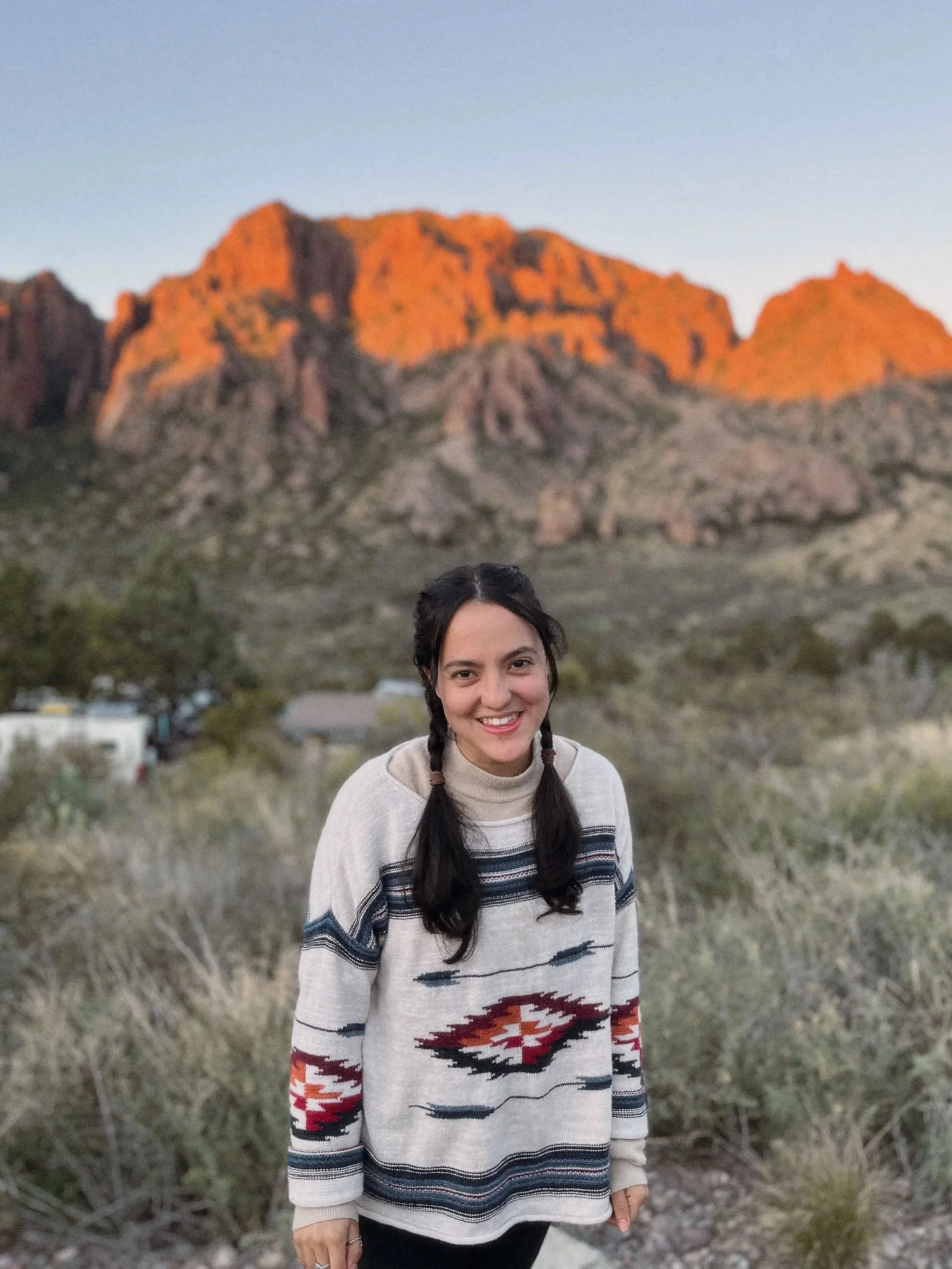 A young woman with long dark hair styled in two braids, wearing a light-colored sweater with a southwestern pattern, standing outdoors at sunset with red rocks in the background and a clear sky.