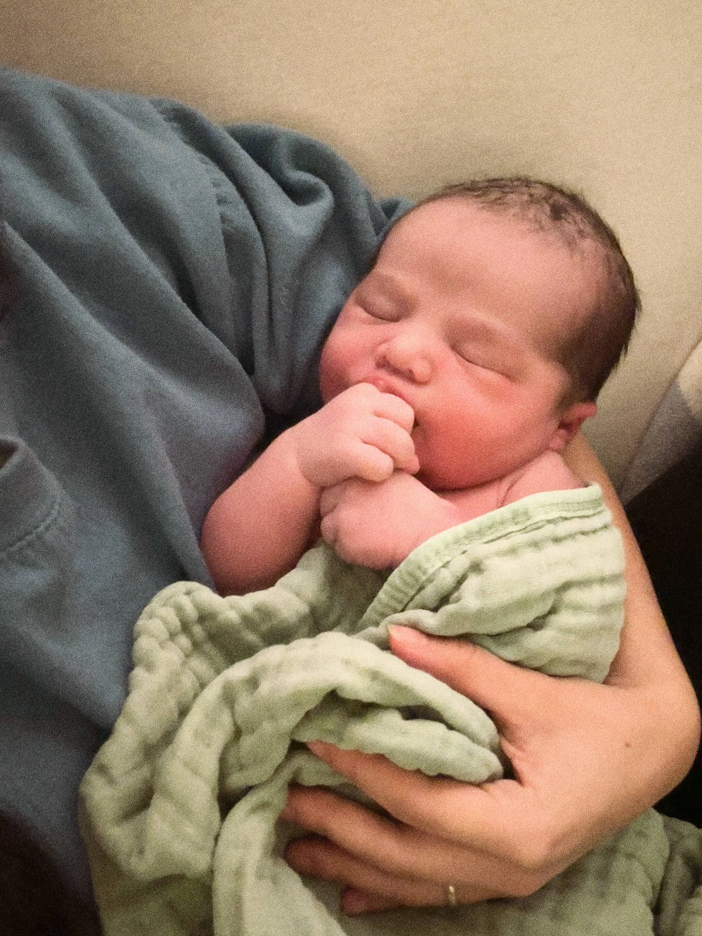 A newborn baby sleeping peacefully, being held gently in someone's arms, wrapped in a soft, green blanket.