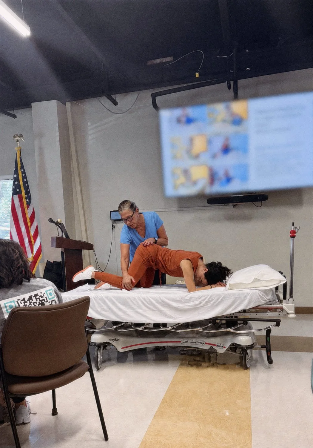 Instructor demonstrating physical therapy techniques on a patient lying face down on a hospital bed in a classroom setting, with an American flag in the background.