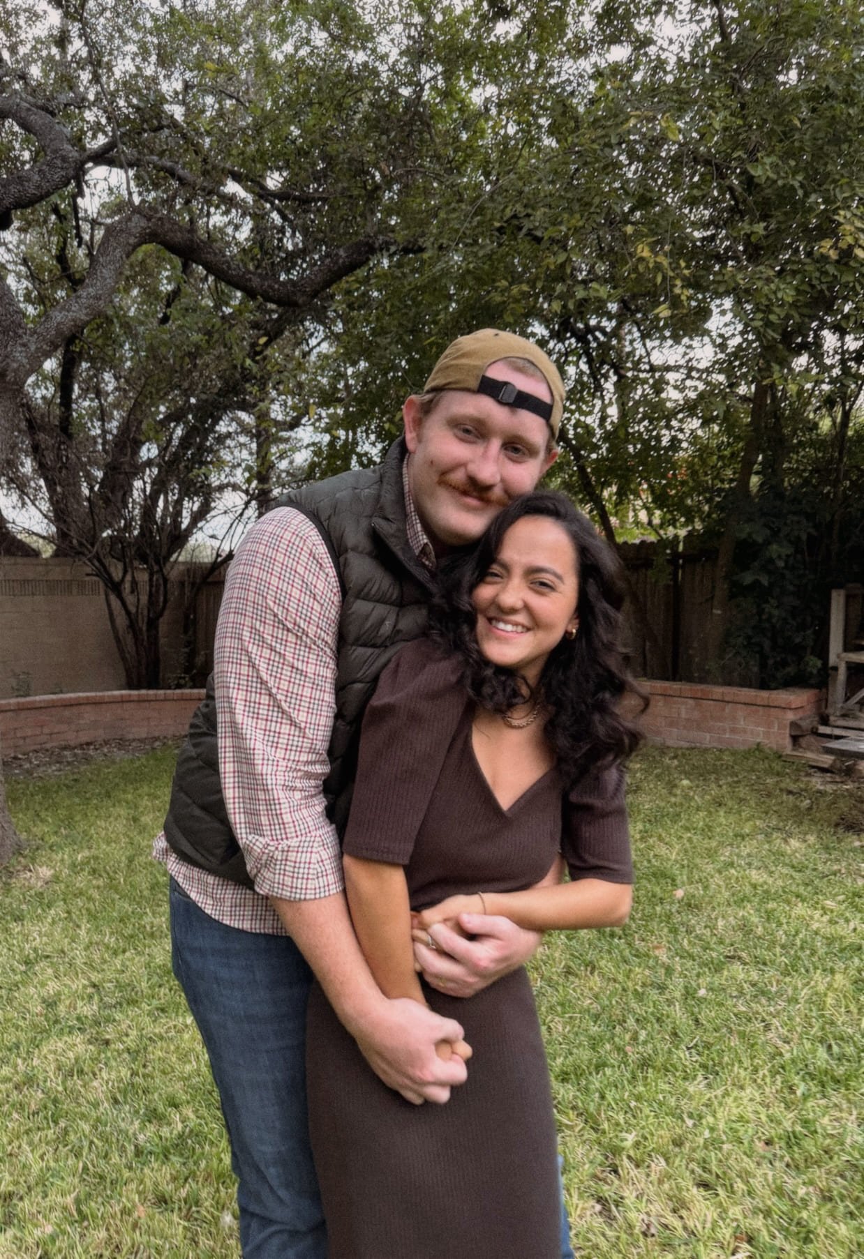 A man and woman standing close together outdoors on a grassy area, smiling and embracing. The man wears a beige cap backwards, a gray vest over a plaid shirt, and jeans. The woman has dark curly hair, a dark brown outfit, and is smiling with her eyes closed.