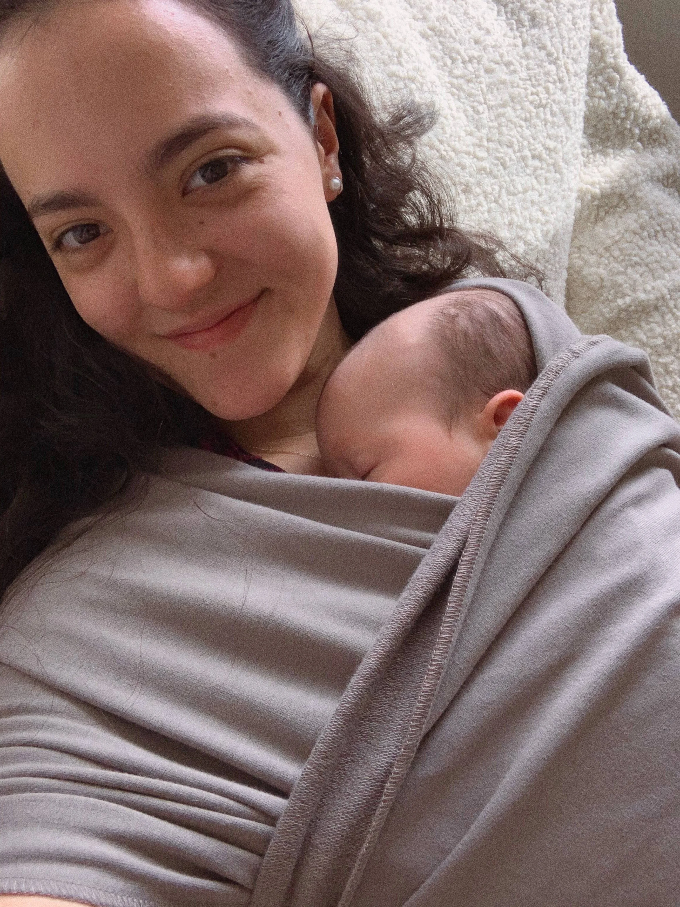 A woman lying down with a baby close to her chest, both resting on a cozy, textured blanket.