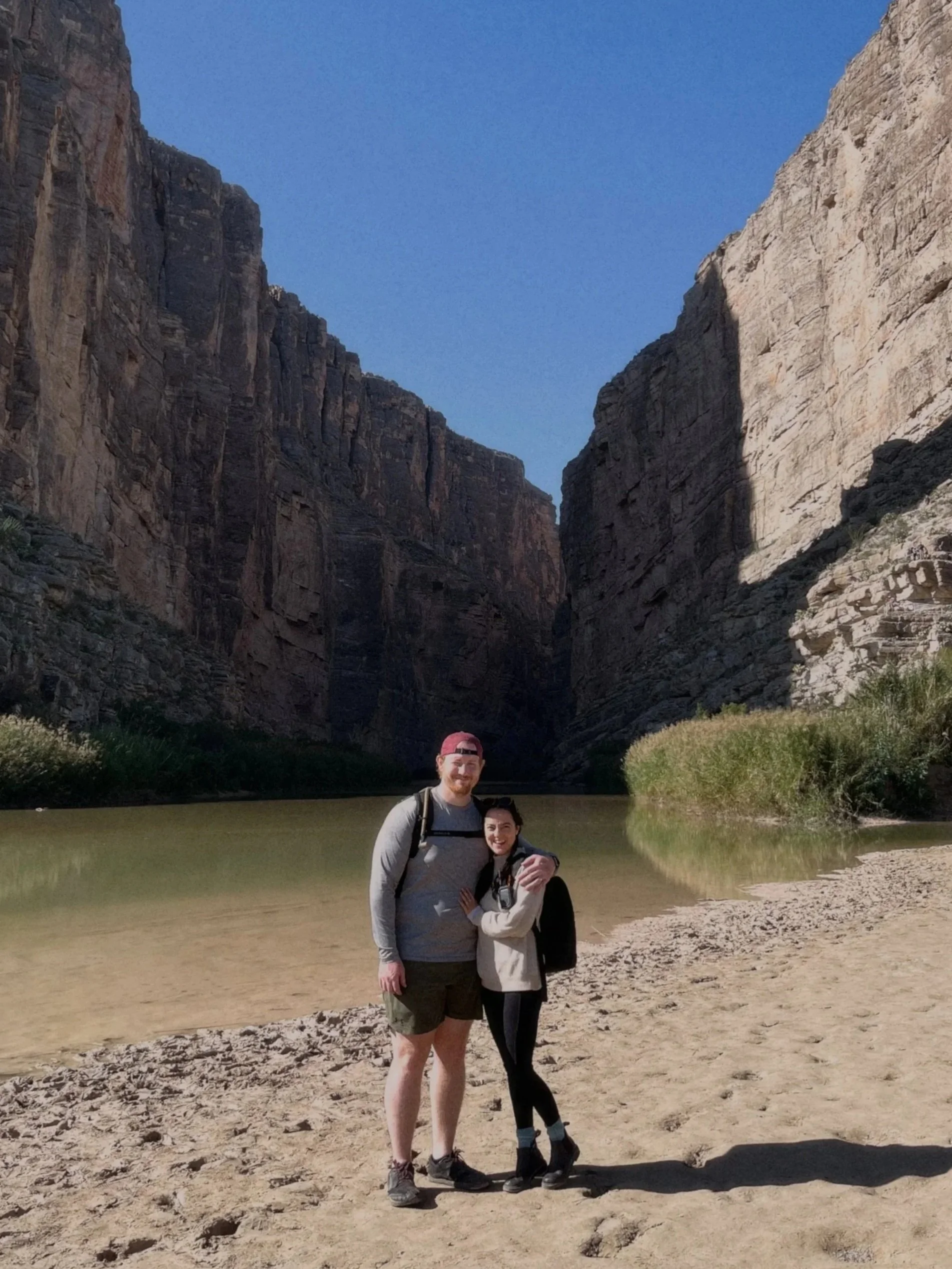 Two people, a man and a woman, standing close together both smiling, in front of a body of water with tall rocky cliffs and clear blue sky in the background.