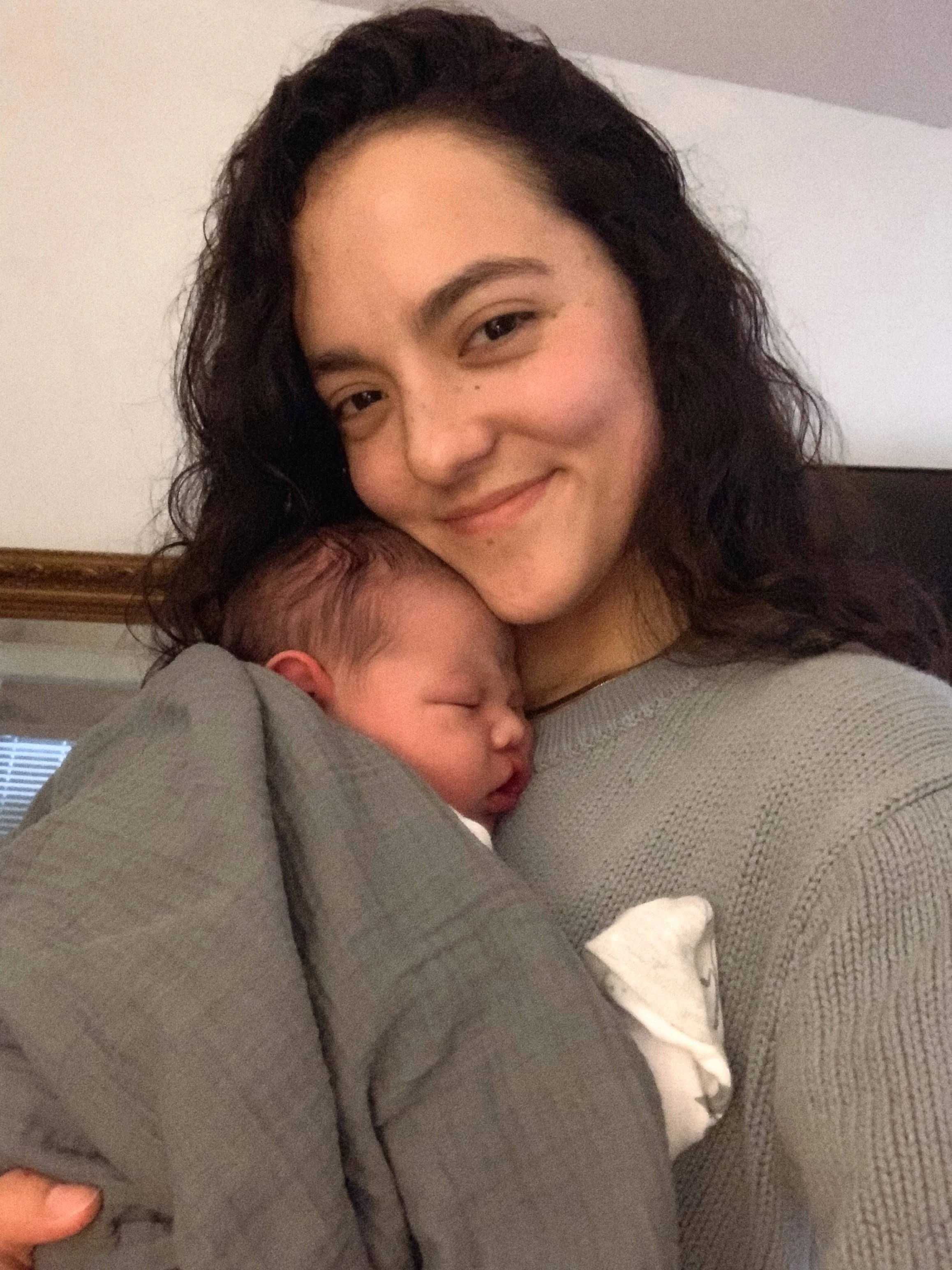 A woman with dark curly hair is smiling and holding a sleeping baby close to her chest. The woman has light skin and is wearing a gray sweater. The background shows part of a wall and a window with blinds.