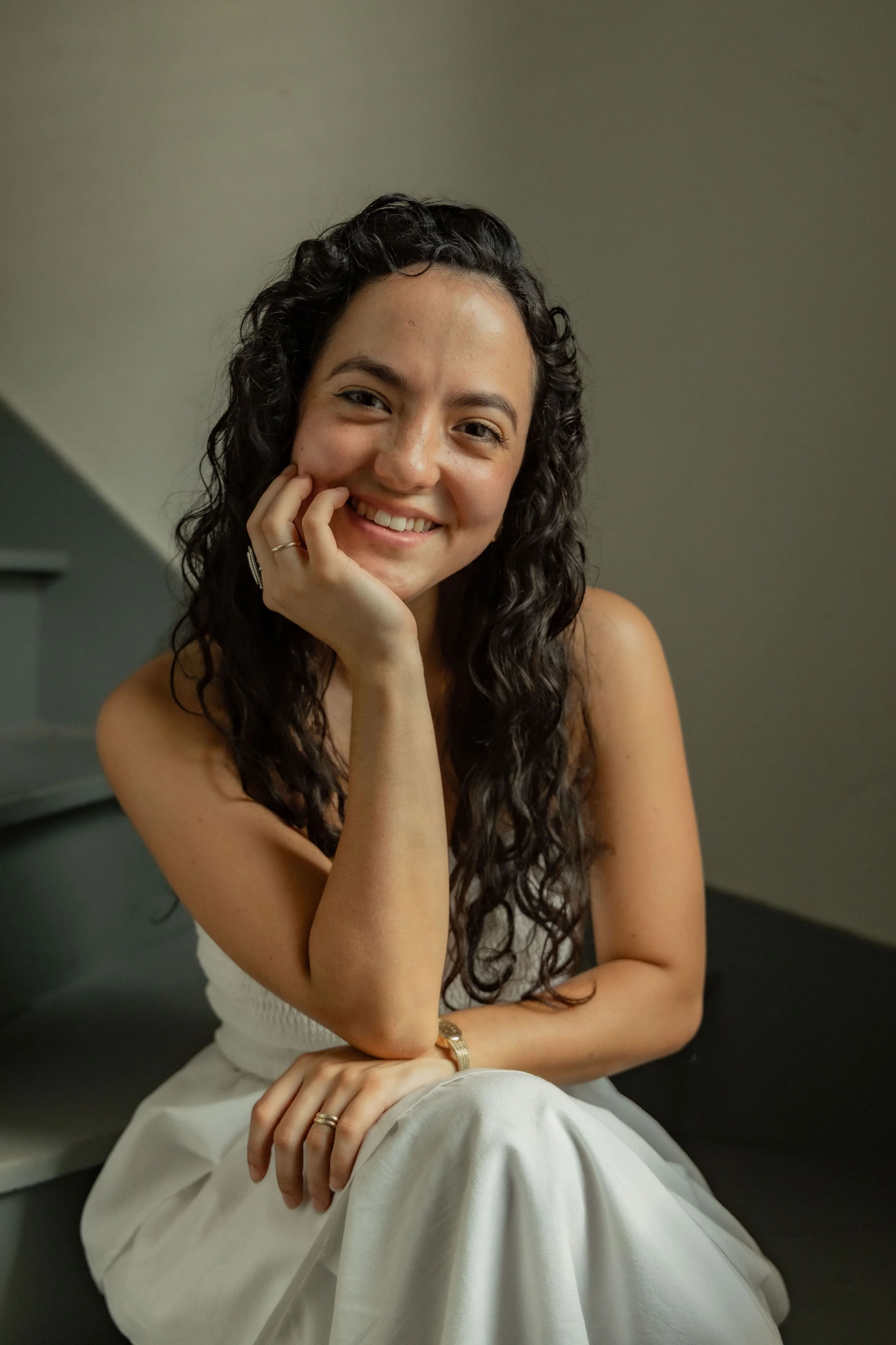 A young woman with curly dark hair, smiling and resting her chin on her hand, wearing a white sleeveless top and white pants, sitting indoors in a relaxed pose.
