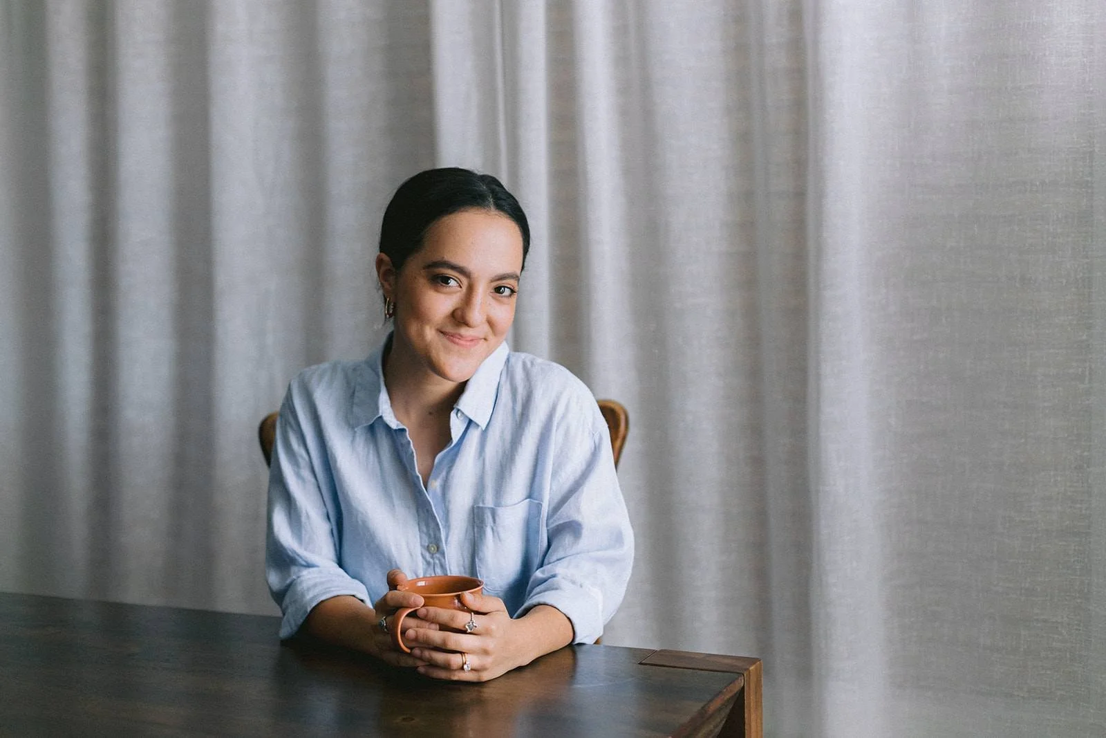 A woman sitting at a dark wooden table, holding a mug with both hands, smiling, wearing a light blue button-up shirt, with a neutral background of gray curtains.