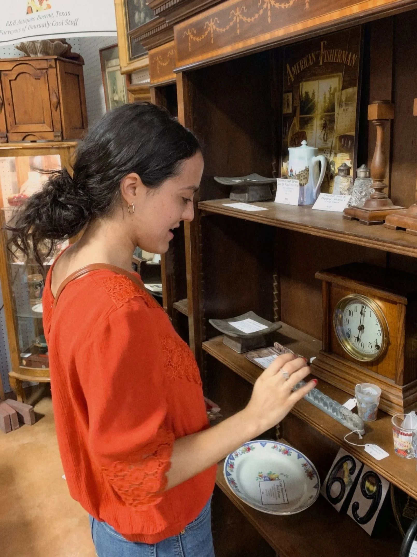 A woman with dark hair in a ponytail, wearing an orange lace top and jeans, shopping for antique items in a store, examining a small object on a wooden shelf.