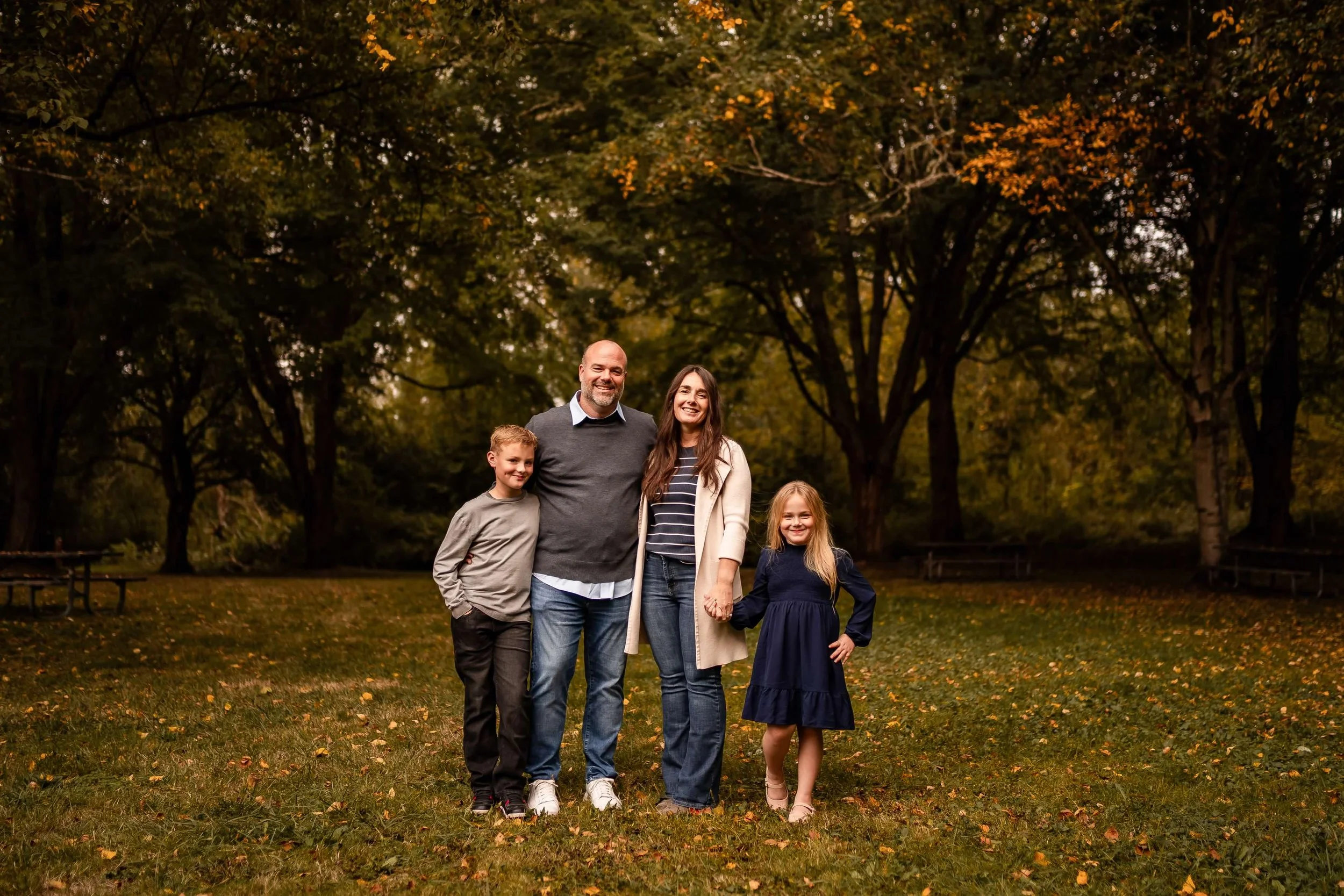 A happy family of five standing on a grassy field in a park during fall, holding hands and smiling, with trees and fallen leaves in the background.