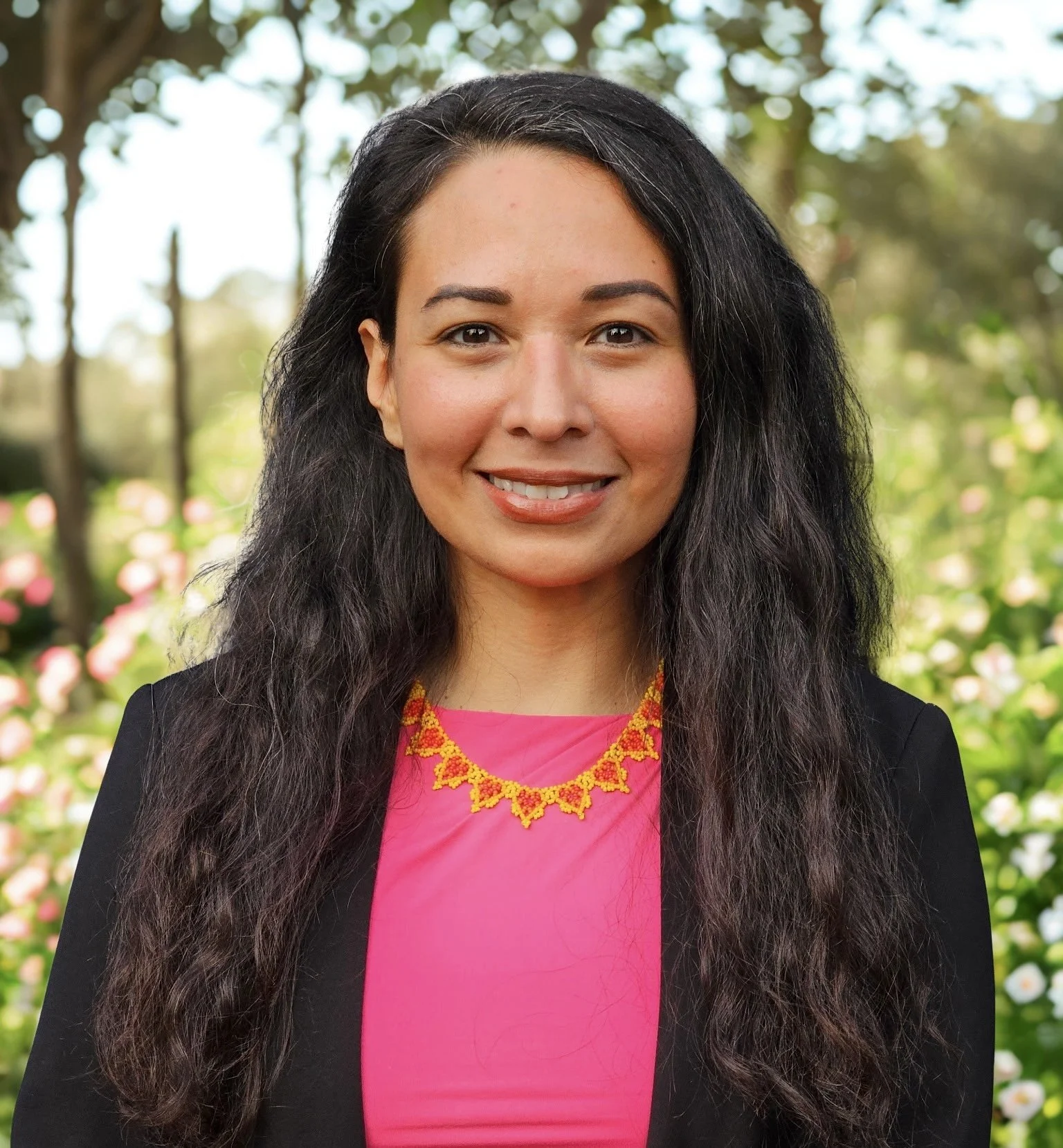 A woman with long black hair wearing a black blazer over a pink top and a colorful beaded necklace, standing outdoors with a background of trees and flowers.