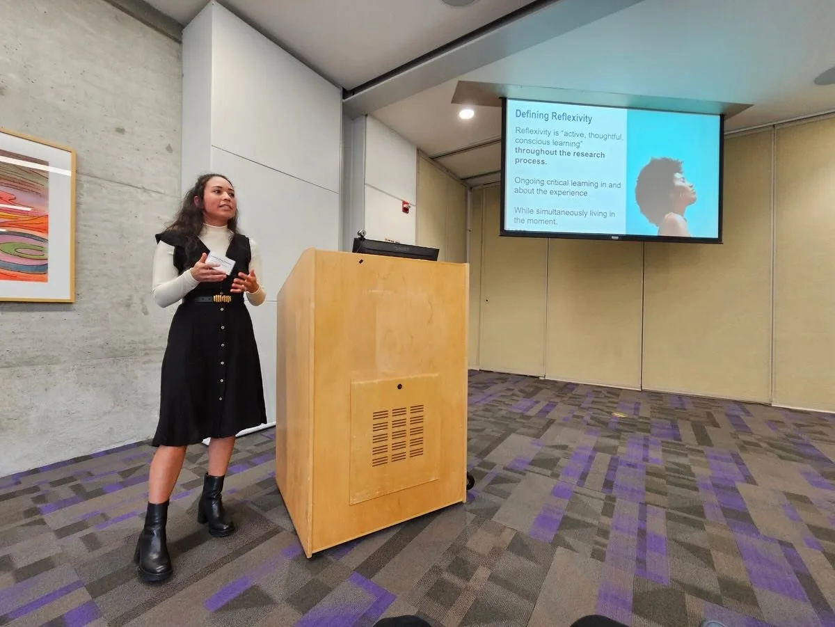 A young woman giving a presentation in a conference room with a large screen displaying a slide titled "Defining Reflexivity." She is standing beside a wooden podium, wearing a black dress, white turtleneck, and black boots, with a piece of paper in her hands.
