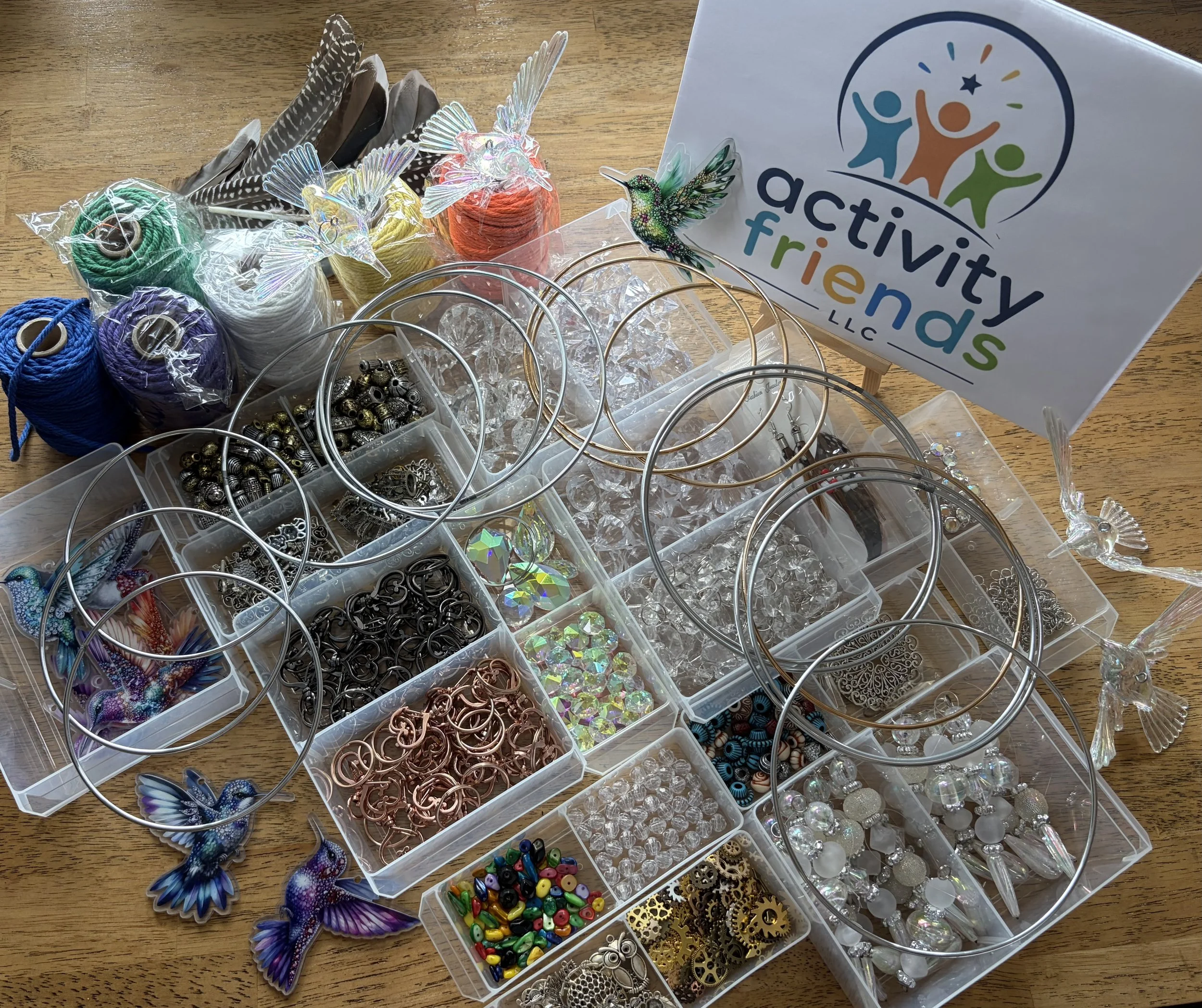 A collection of craft supplies, including colorful yarn balls, various metal and plastic jewelry components, decorative beads, and sticker decorations, arranged on a wooden surface. There is also a sign with the logo of 'Activity Friends LLC' featuring stylized human figures and bright colors.
