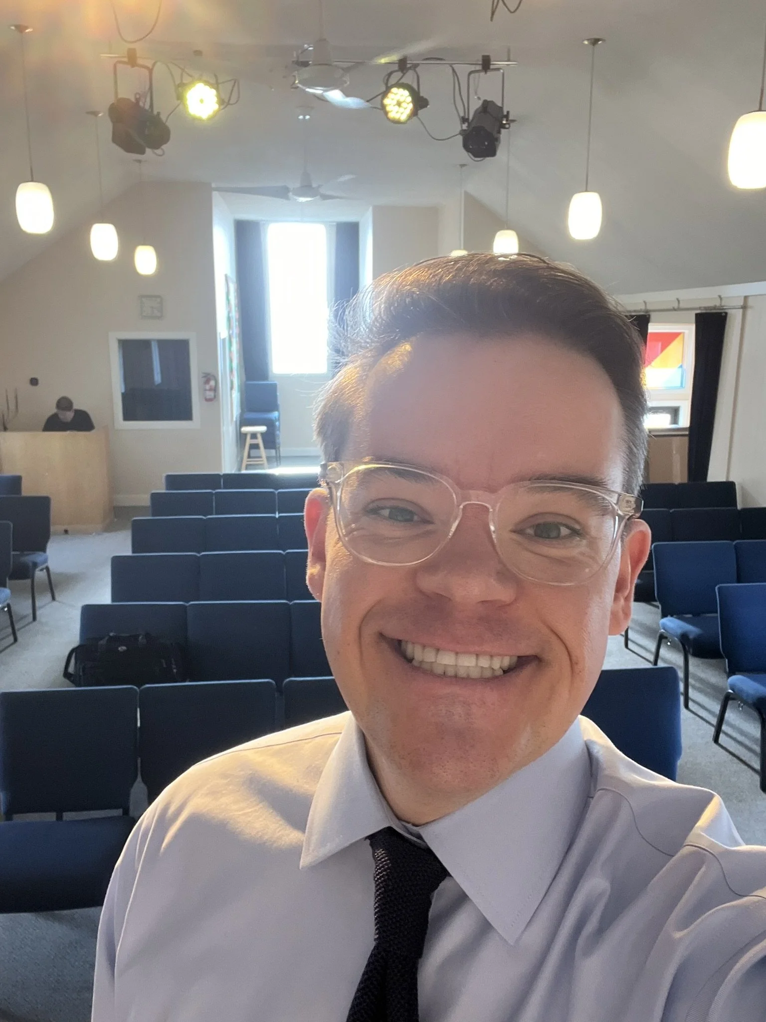 A smiling man in a white shirt and black tie taking a selfie inside a large, well-lit conference room with blue chairs arranged in rows.