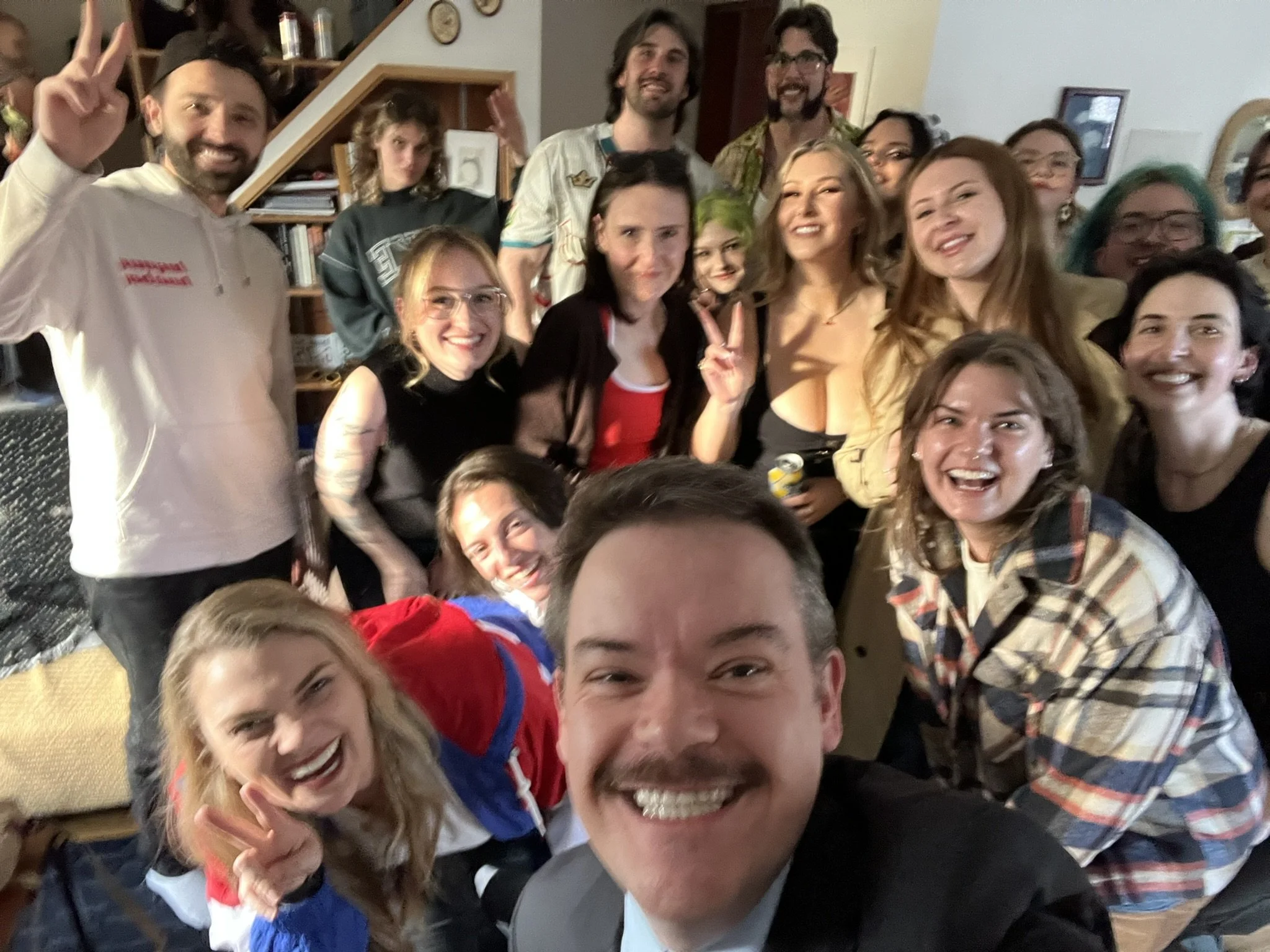 Group of people smiling and posing for a photo at a gathering or party, with some making peace signs.