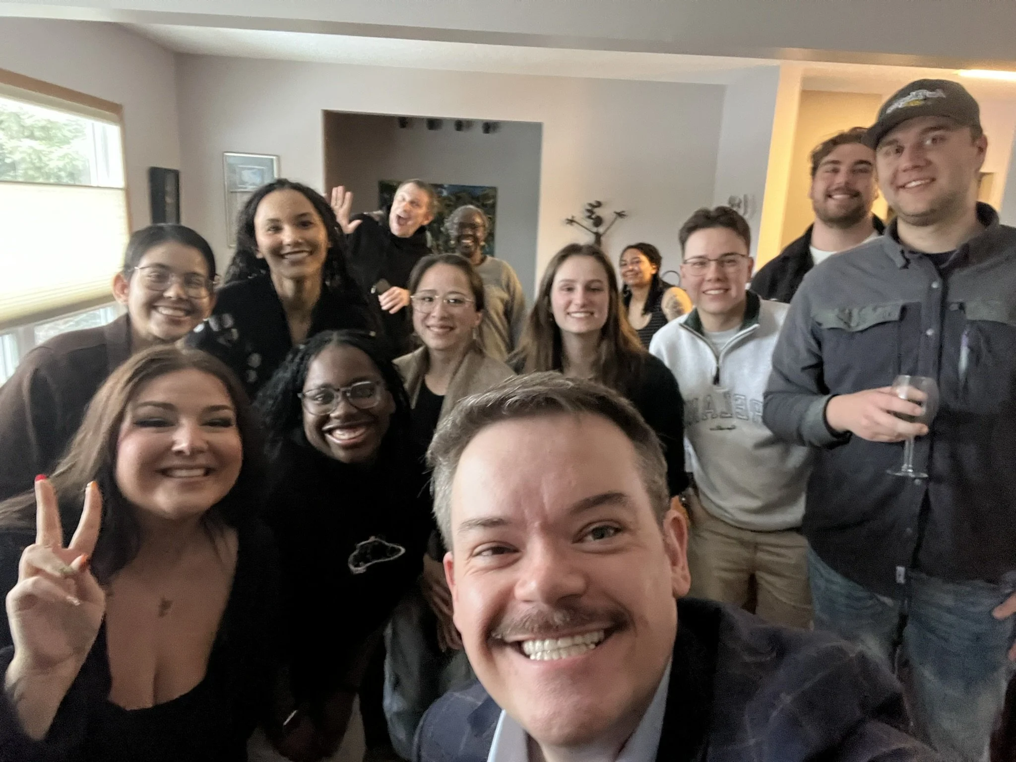 Group of smiling people gathered indoors, some making peace signs, with a person in front taking a selfie, during a social gathering or celebration.