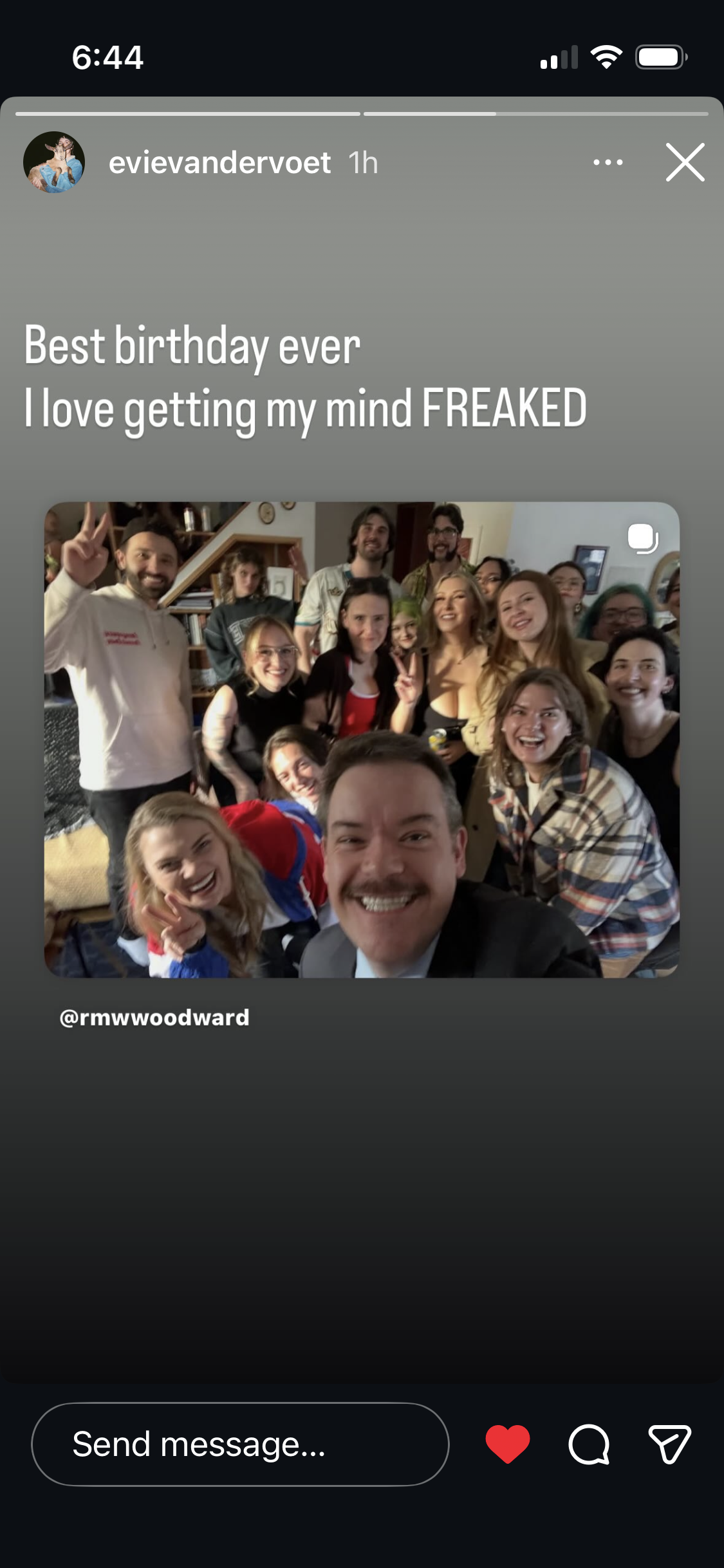 A group of people smiling and posing for a selfie in a living room, with a staircase and bookshelf in the background. Most are making peace signs or smiling broadly.