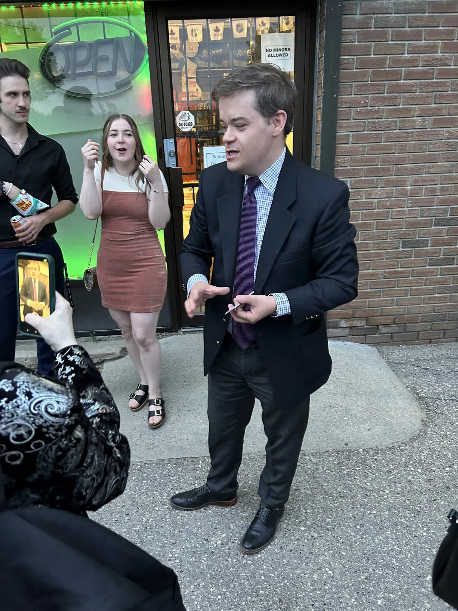 A man in a dark suit and purple tie is signing autographs for fans outside a store with a brick wall. Behind him, a young woman in a brown dress and sandals, and a young man holding snacks are visible. Someone is taking a photo with a smartphone. The