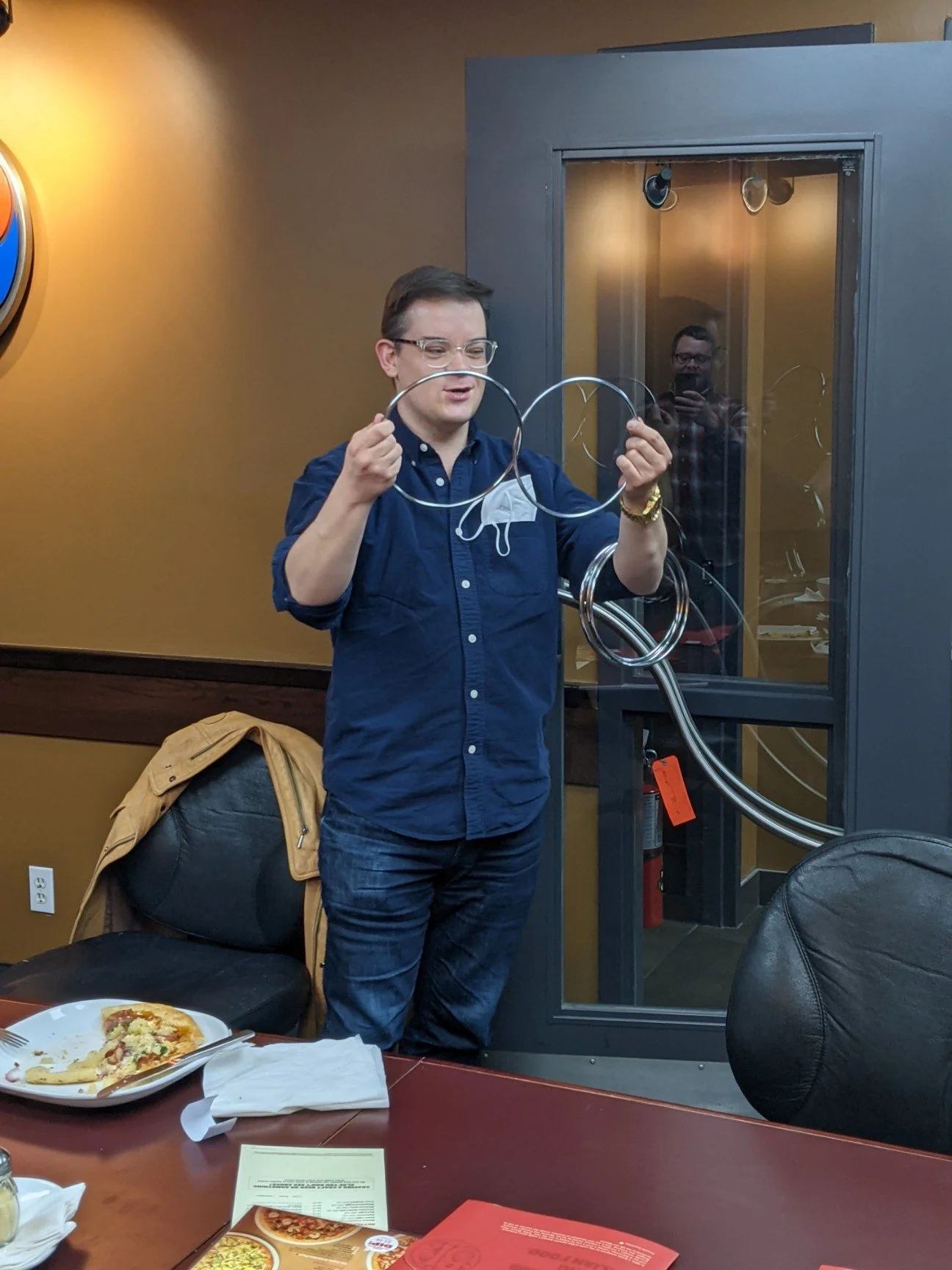 A man with glasses and a navy shirt stands in a room with brown and gray walls, holding several metal hoops. In the background, a person is taking a photo through a glass door. In the foreground, there is a pizza on the table, with a yellow jacket dr