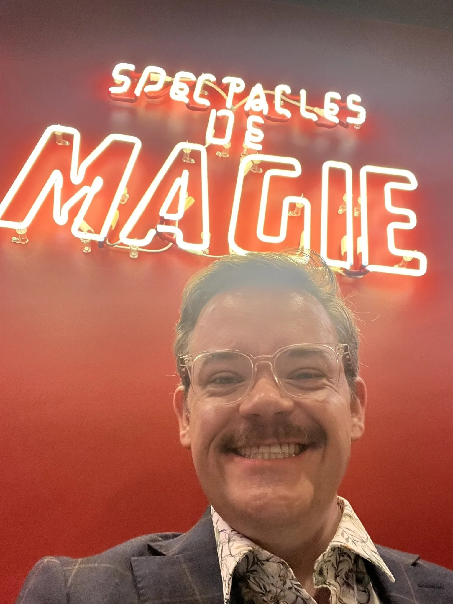 A man smiling, wearing glasses, a dark plaid blazer, and a patterned shirt, standing in front of a red wall with a neon sign that reads 'Spectacles' at Le Double Fond magician's club in Paris. 