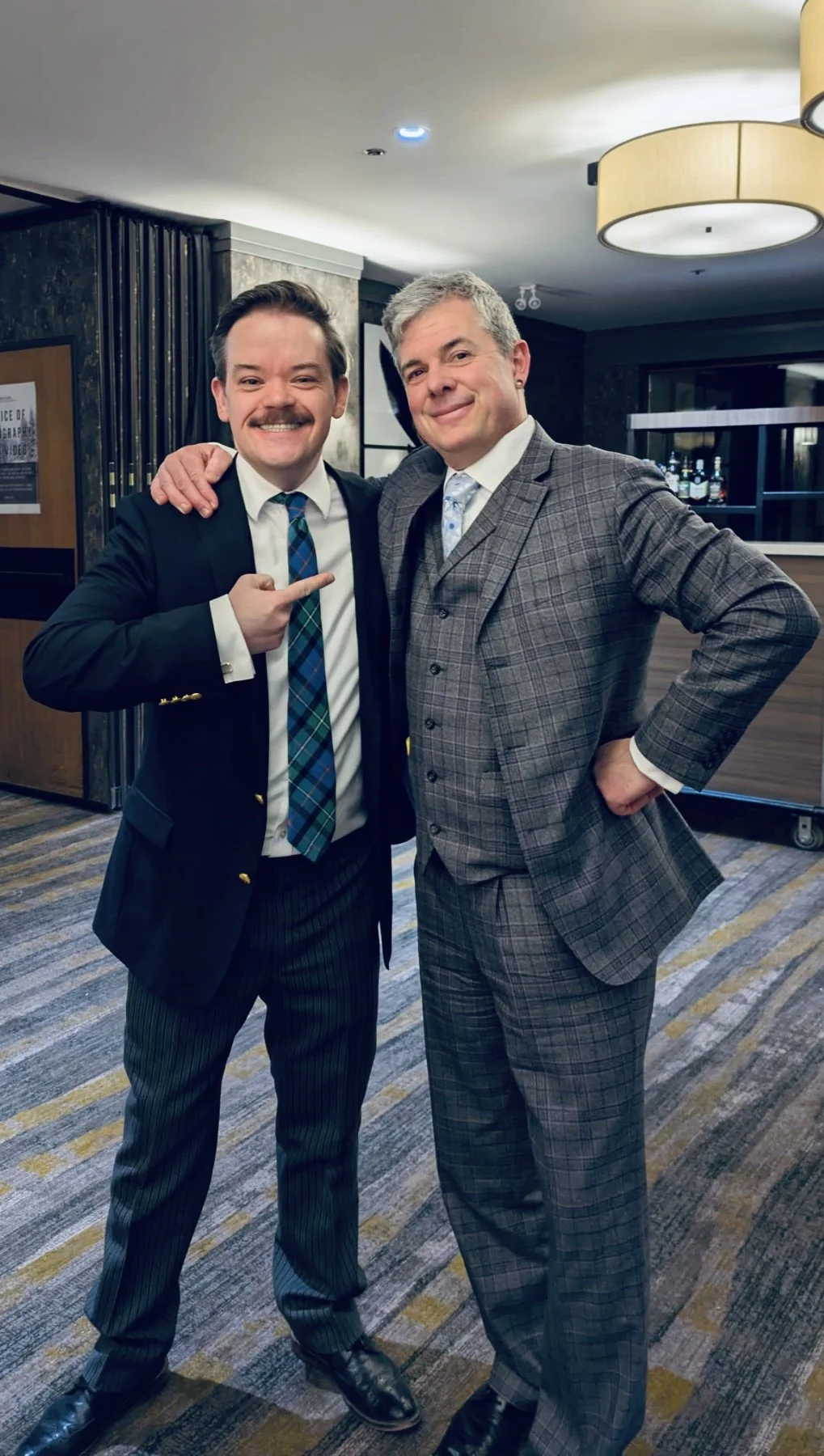 Two men in suits standing closely together indoors, smiling at the camera. One man on the left is pointing at the other with an arm around his shoulder. The man on the right has his hand on his hip. The background includes a bar area and modern decor