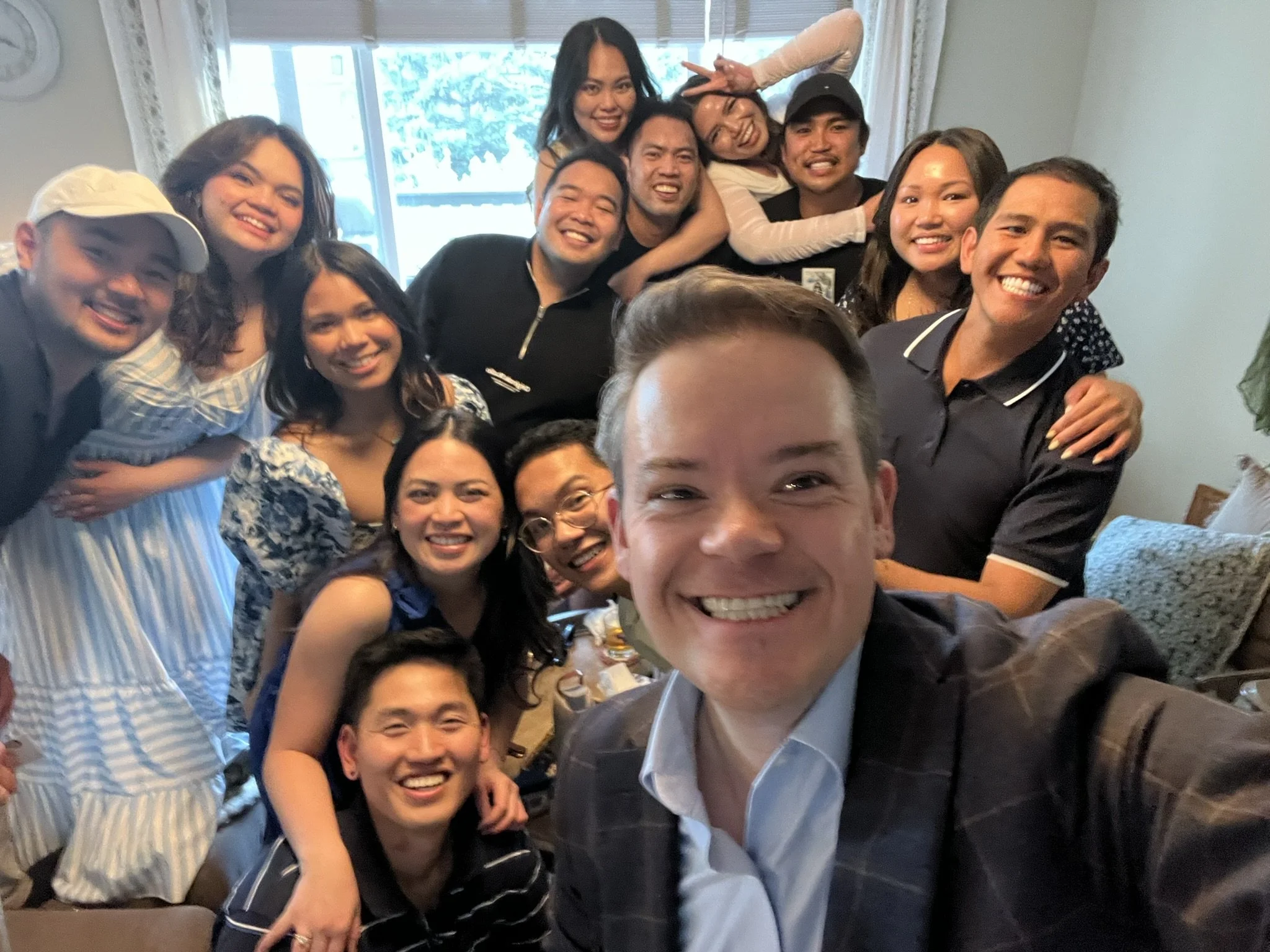 A group of people smiling and posing closely together for a photo indoors, with a window and curtain visible in the background.