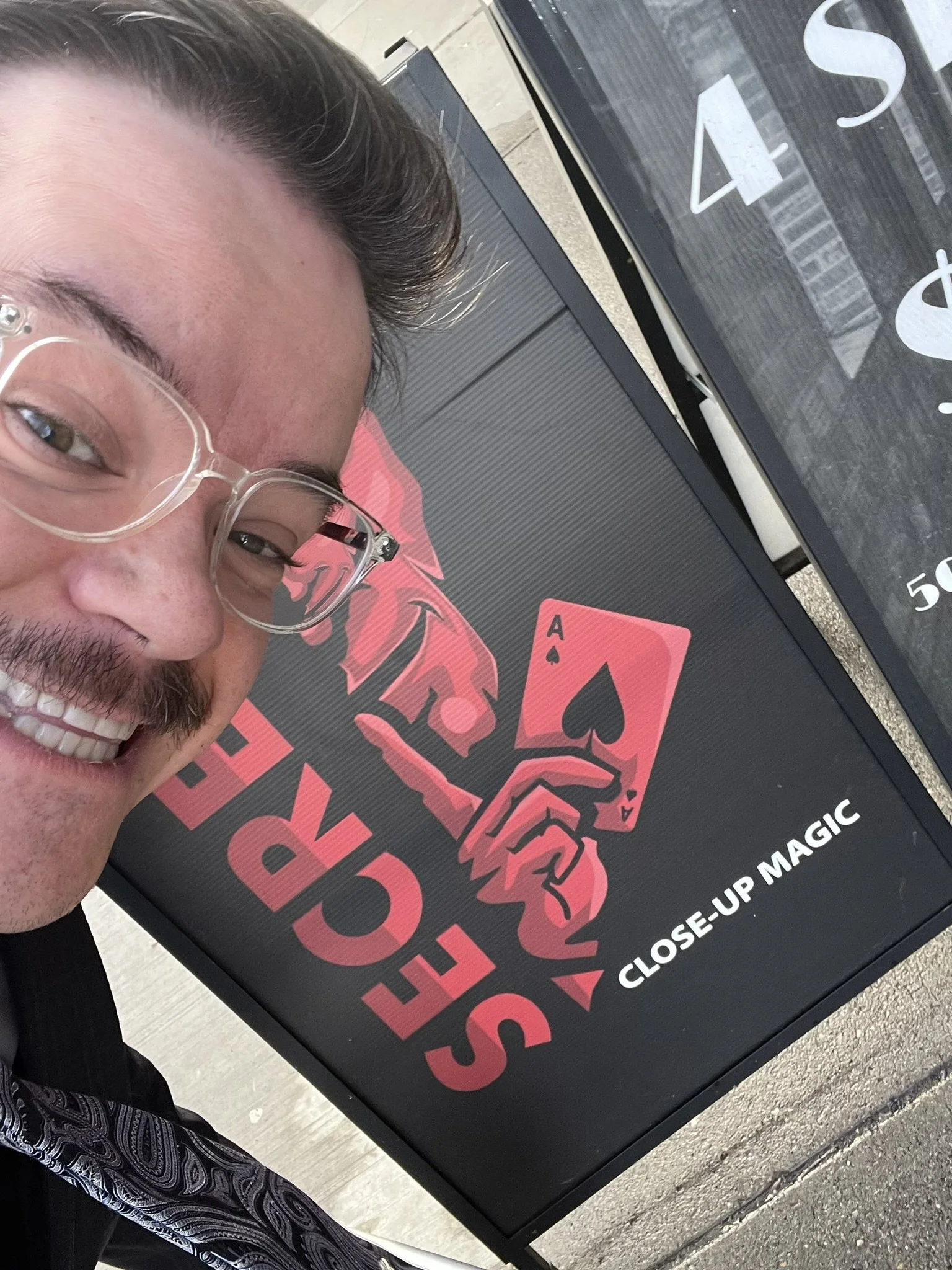 Close-up of a smiling man with glasses taking a selfie in front of a black sign with pink and white text, and playing cards on the ground.